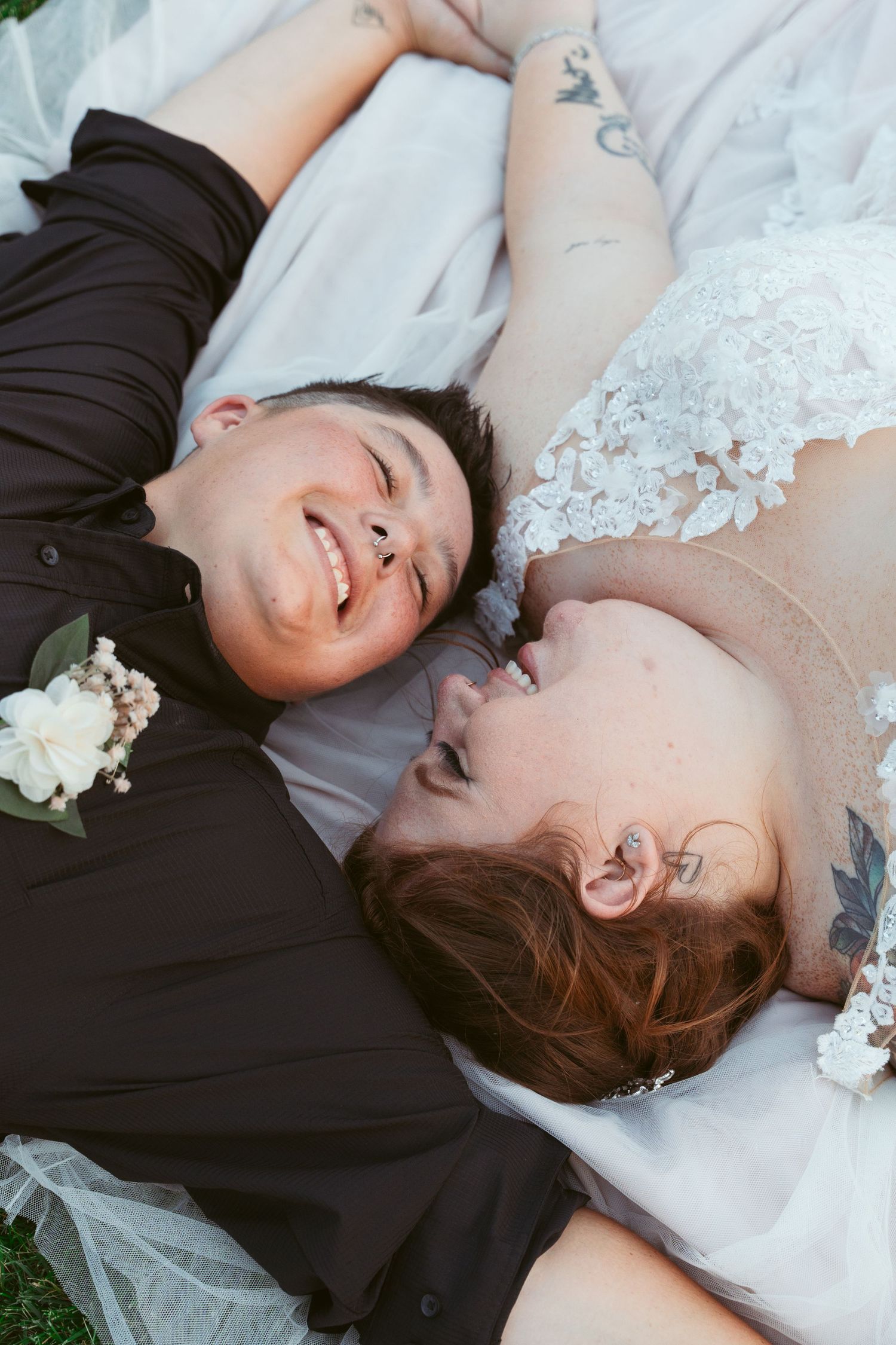 A newlywed couple lying on grass sharing a joyful moment in their wedding attire.