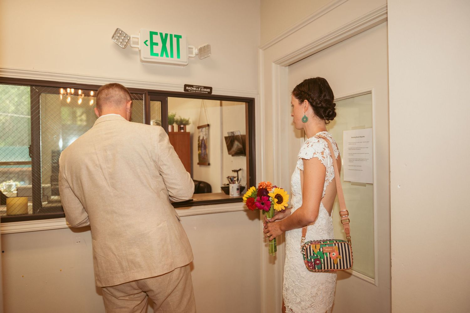 A person in beige clothing holds a colorful bouquet behind their back while walking through a hallway with an exit sign.