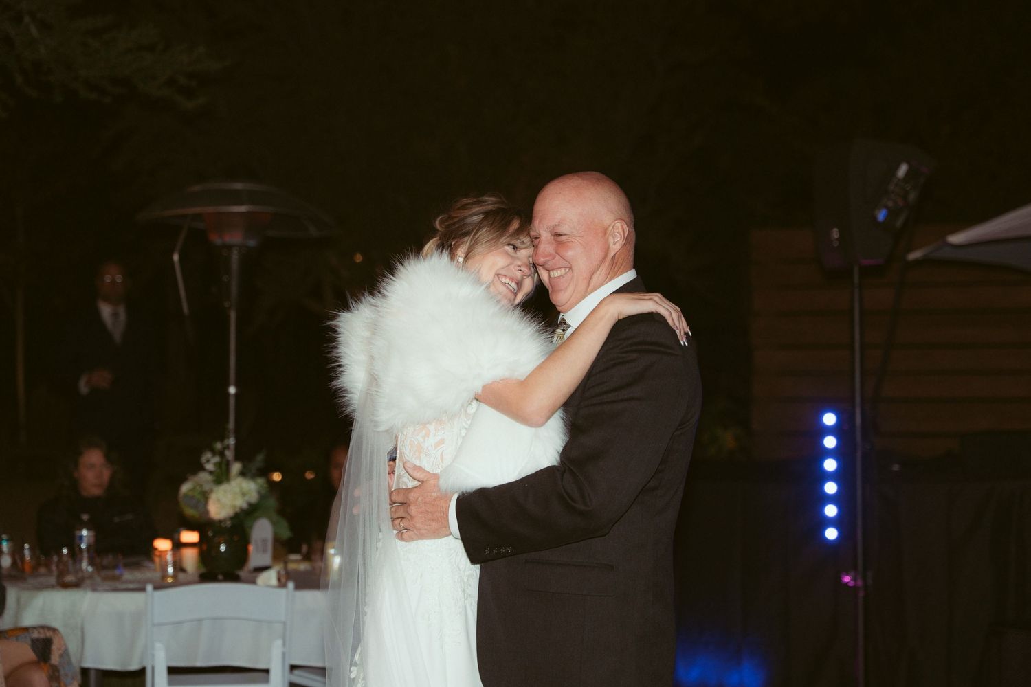 A couple embraces during their wedding dance as guests look on from decorated tables.
