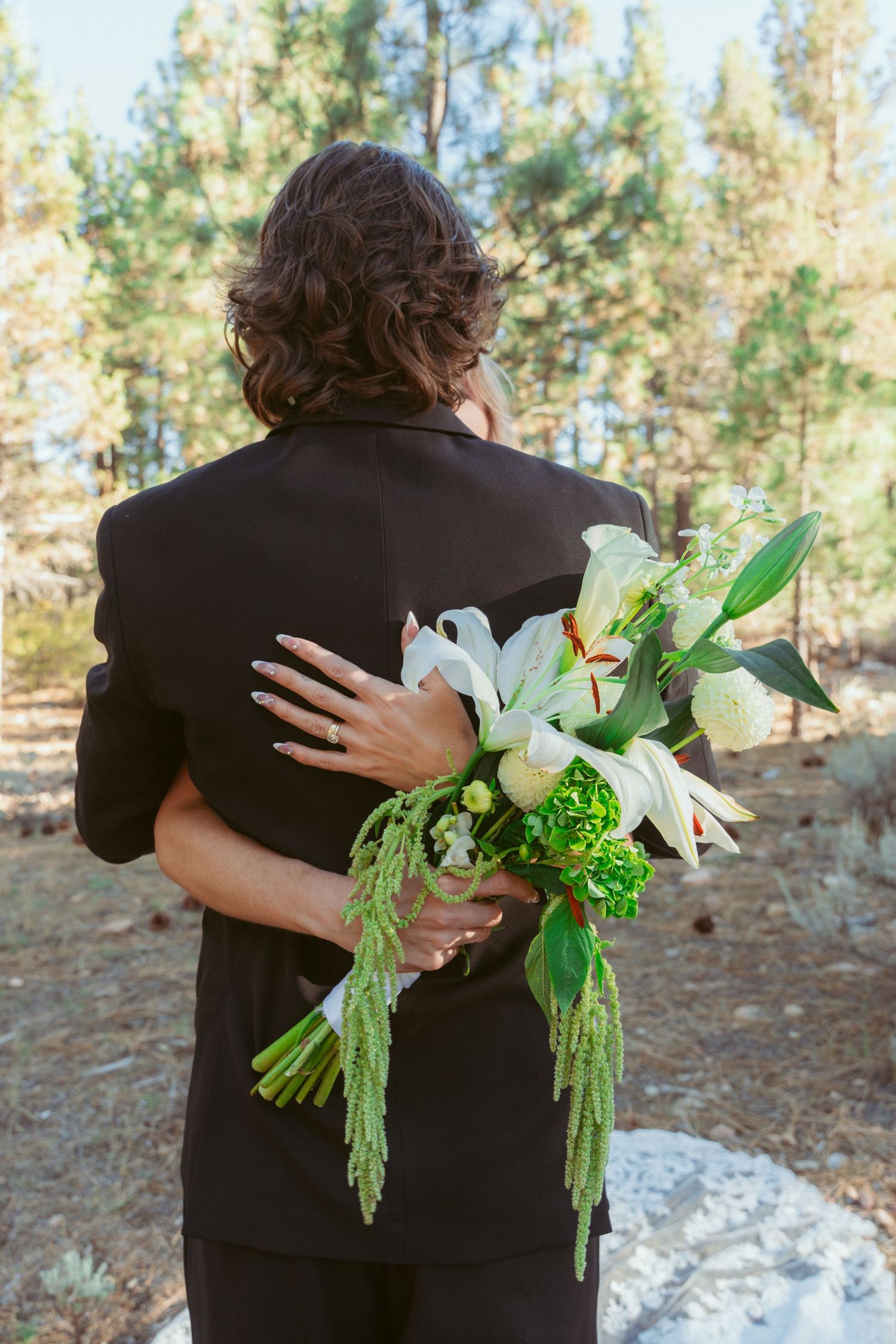 A close-up of white lilies and green foliage in a wedding bouquet against a black suit.