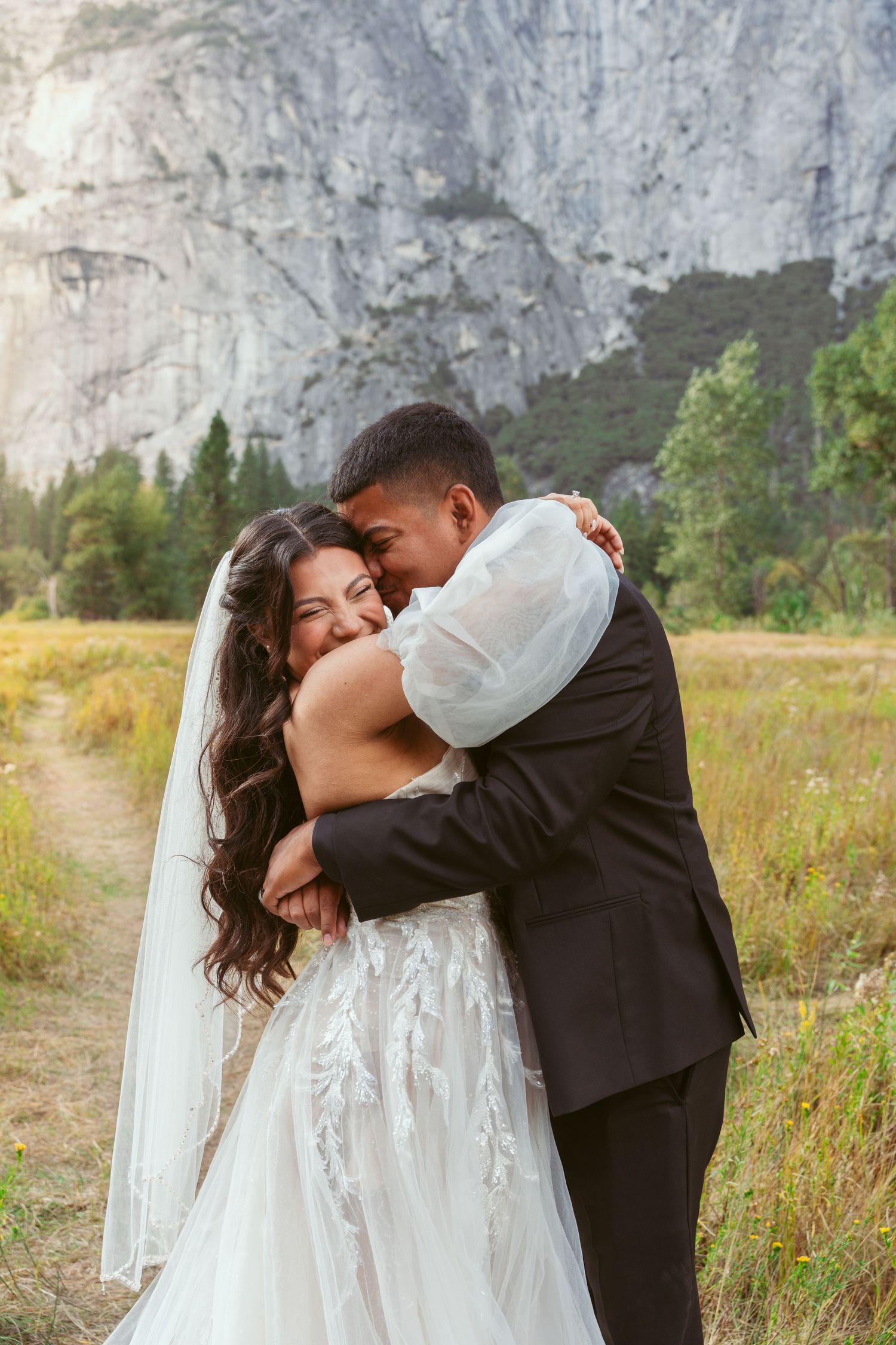 Newlyweds embrace and kiss in a meadow with towering granite cliffs of Yosemite Valley in the background.