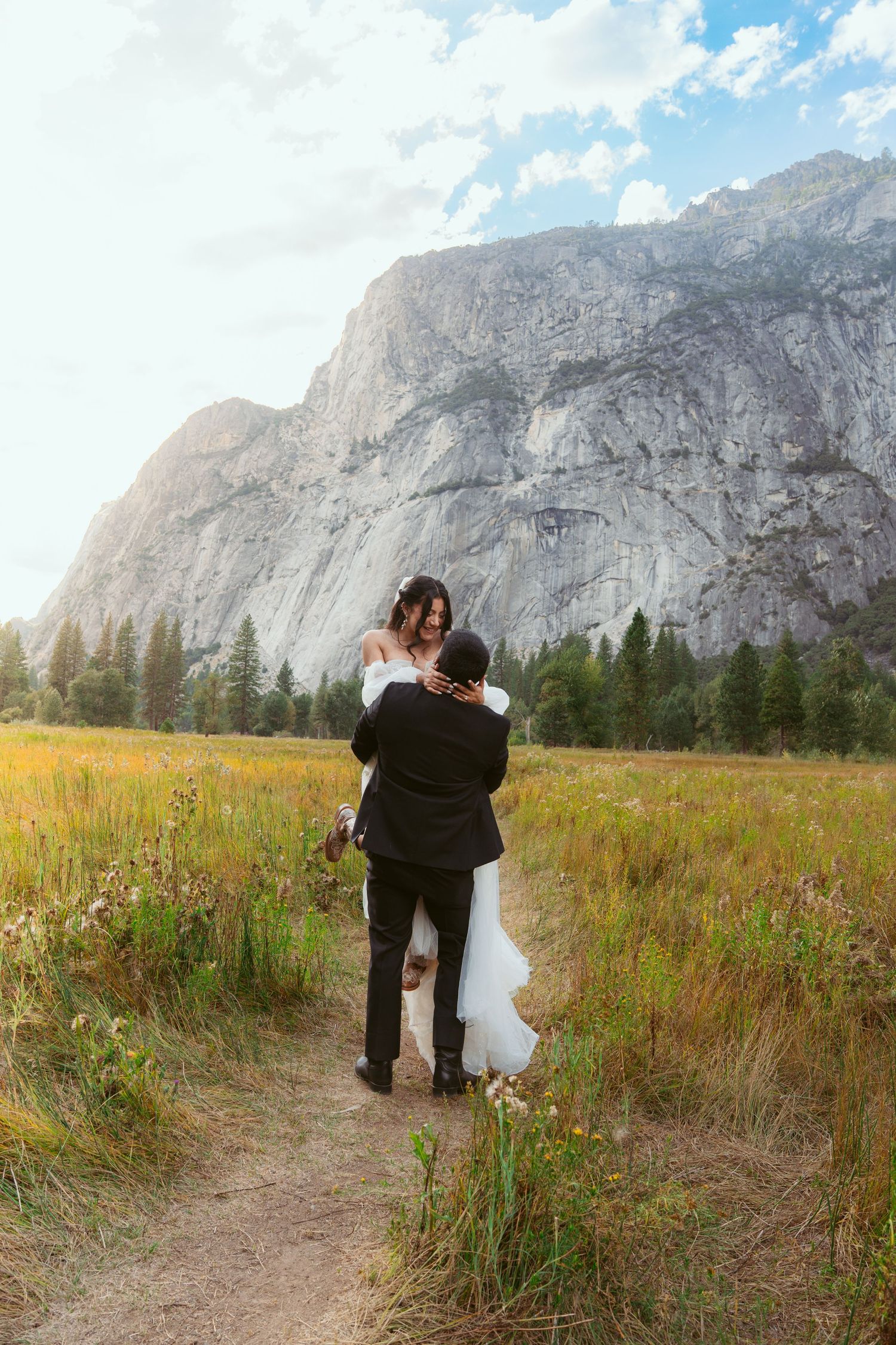 Couple in wedding attire dancing in a wildflower meadow beneath towering mountain cliffs.