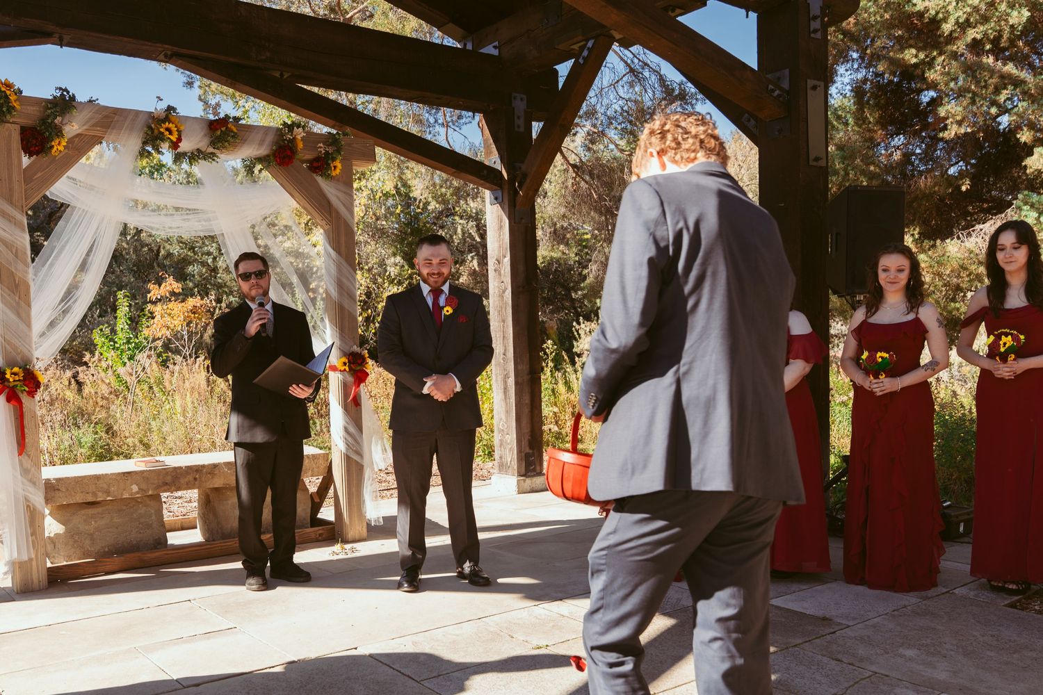 Wedding ceremony participants gather under a decorated wooden pergola with autumn scenery.