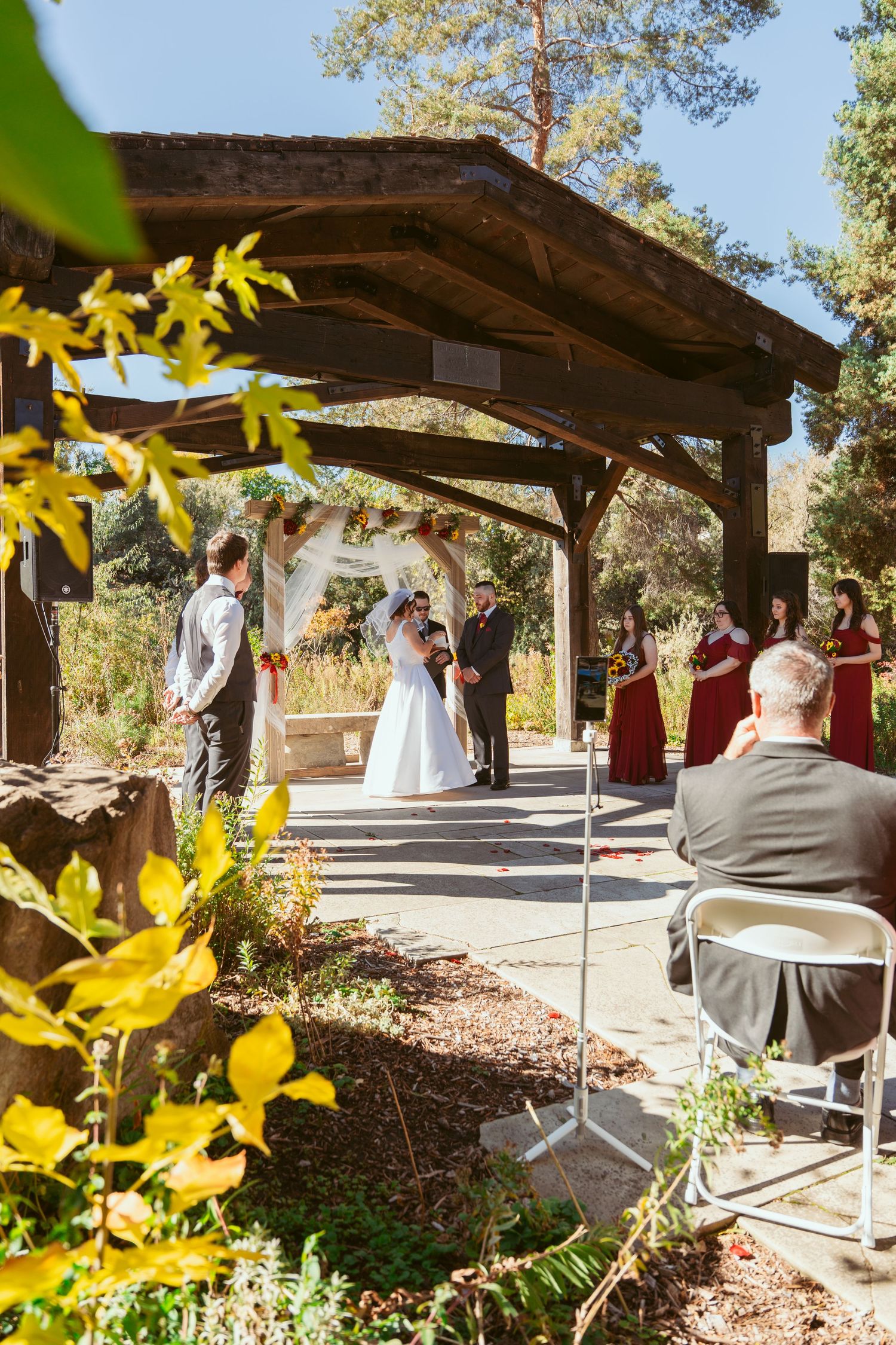 Wedding ceremony view through yellow autumn foliage under rustic wooden pavilion.
