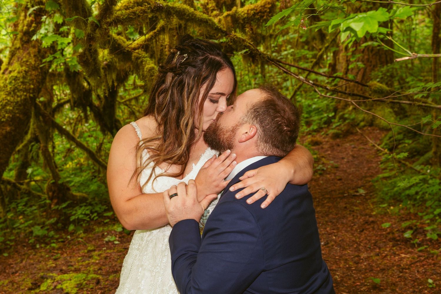 A couple shares an intimate moment among tall trees on a forest path.