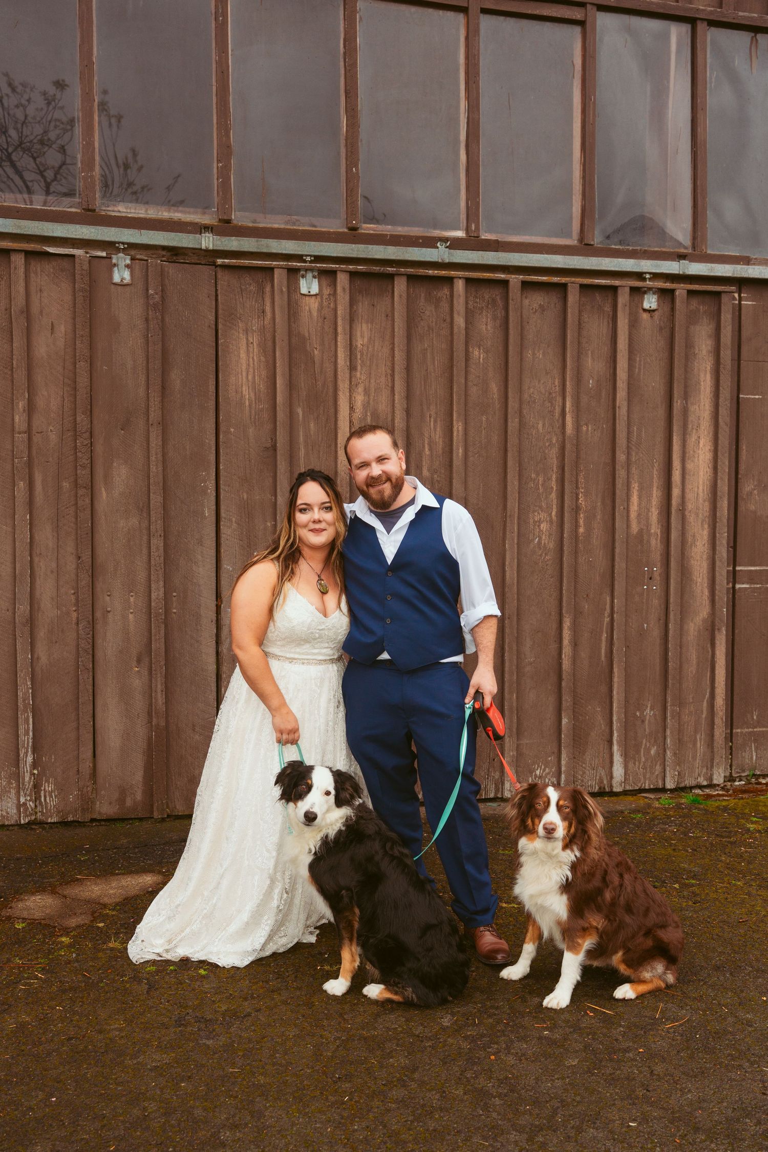 Couple poses with their Australian Shepherd dogs against wooden barn backdrop.