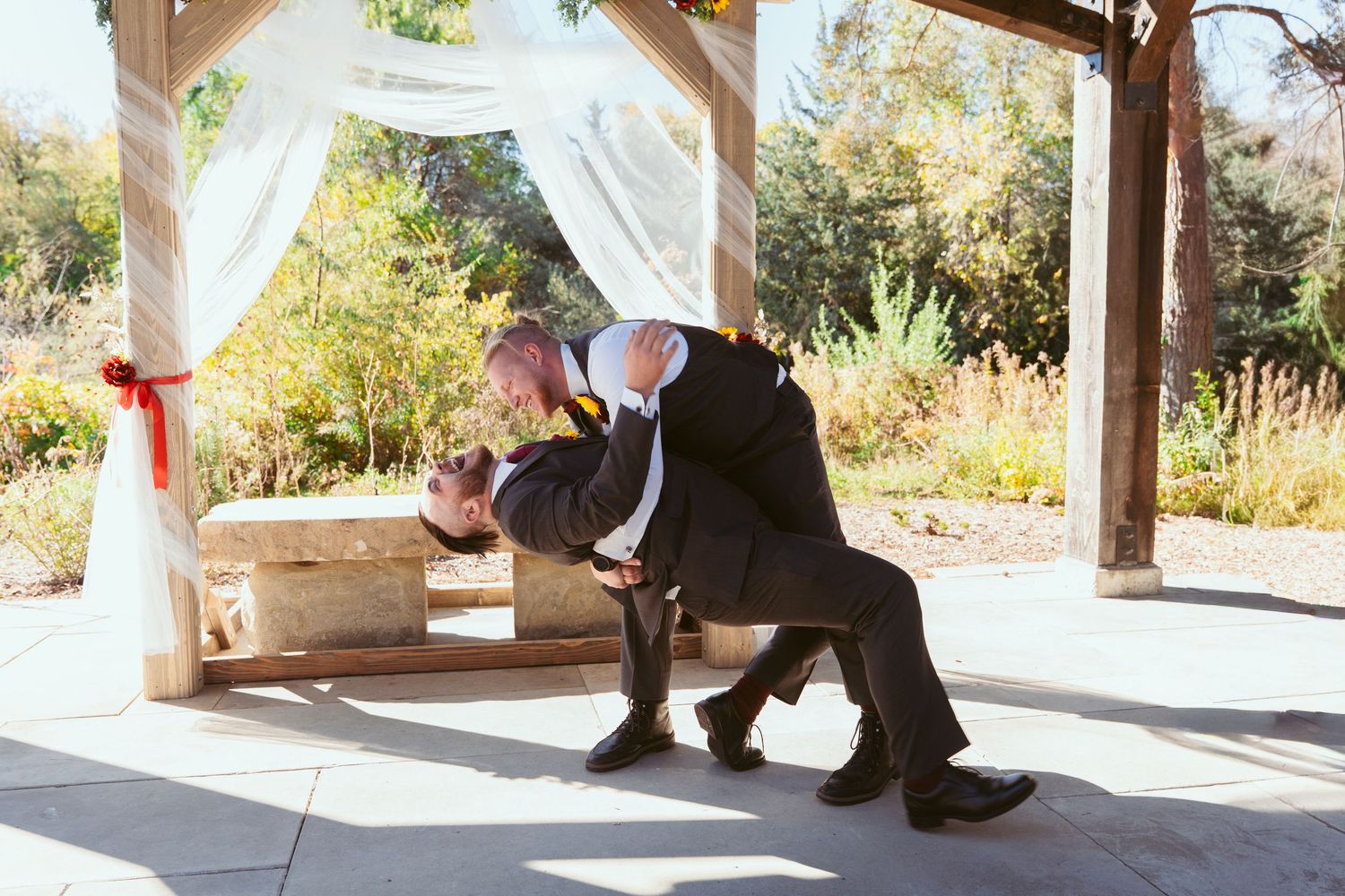 Wedding couple poses under a white draped pergola decorated with red accents and sunflower bouquets.