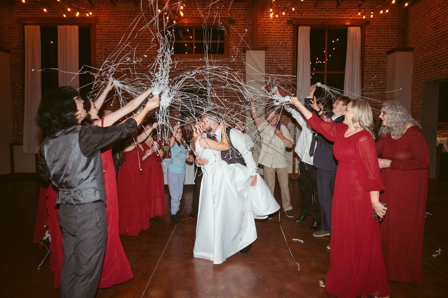 Wedding celebration with guests throwing streamers in a brick-walled venue with string lights.
