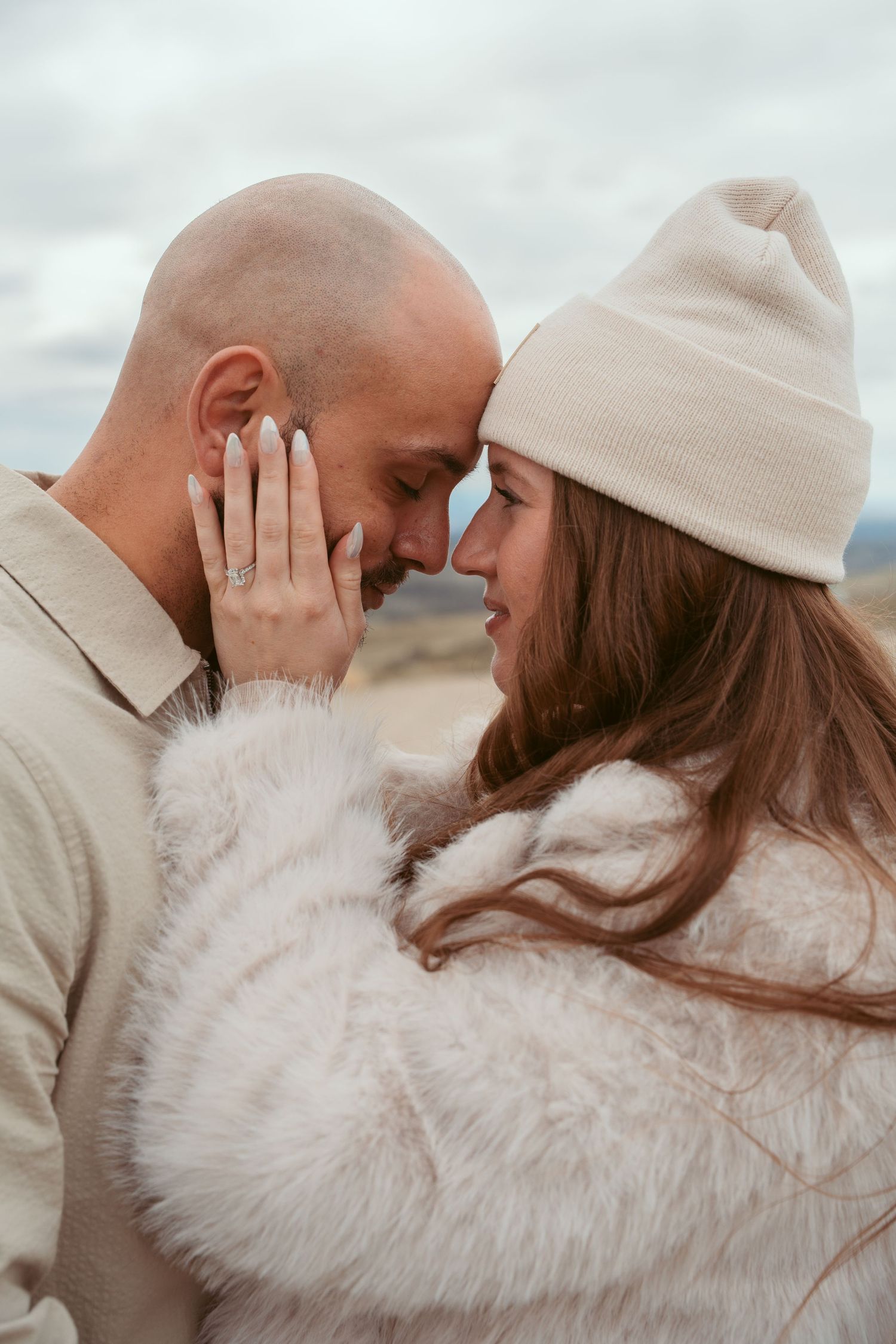 Intimate couple shares tender moment while wearing neutral tones against cloudy backdrop.