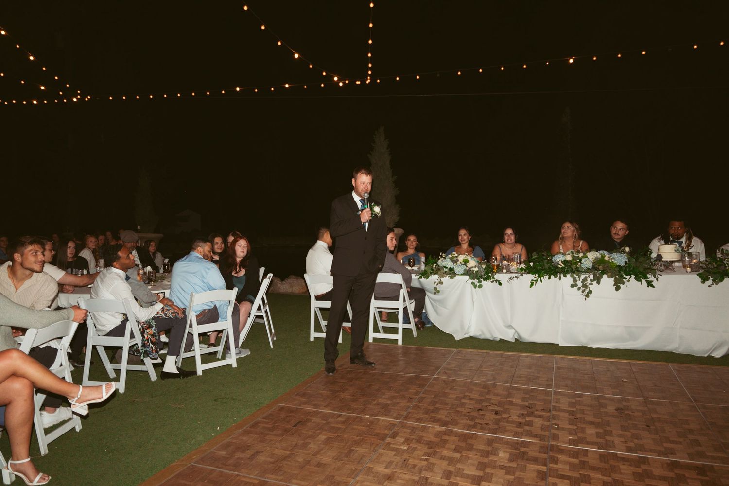Wedding guests enjoying dinner at outdoor reception with string lights overhead and white table settings.