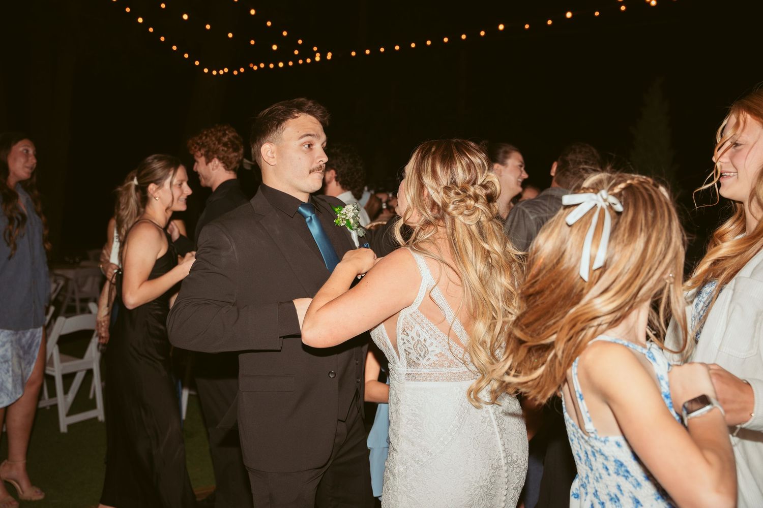 Wedding guests dance and celebrate under strands of twinkling lights at nighttime reception.