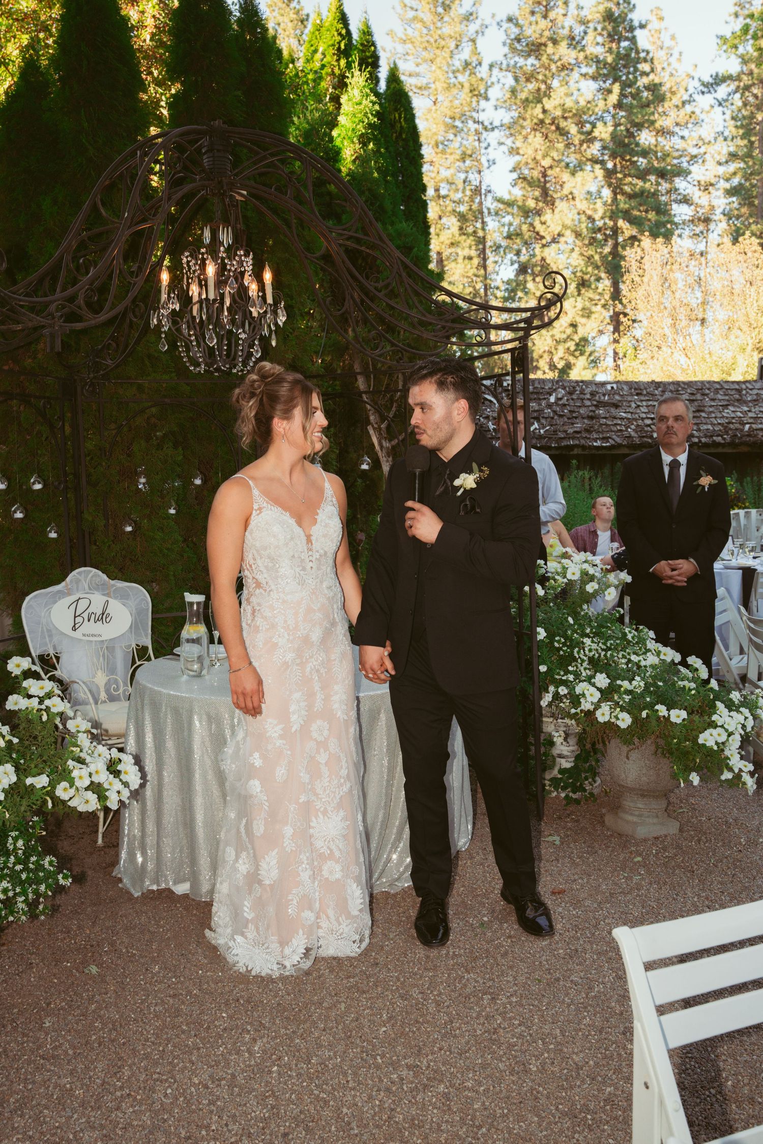 Couple sharing a romantic moment on a garden path surrounded by white floral decorations and evergreen trees.