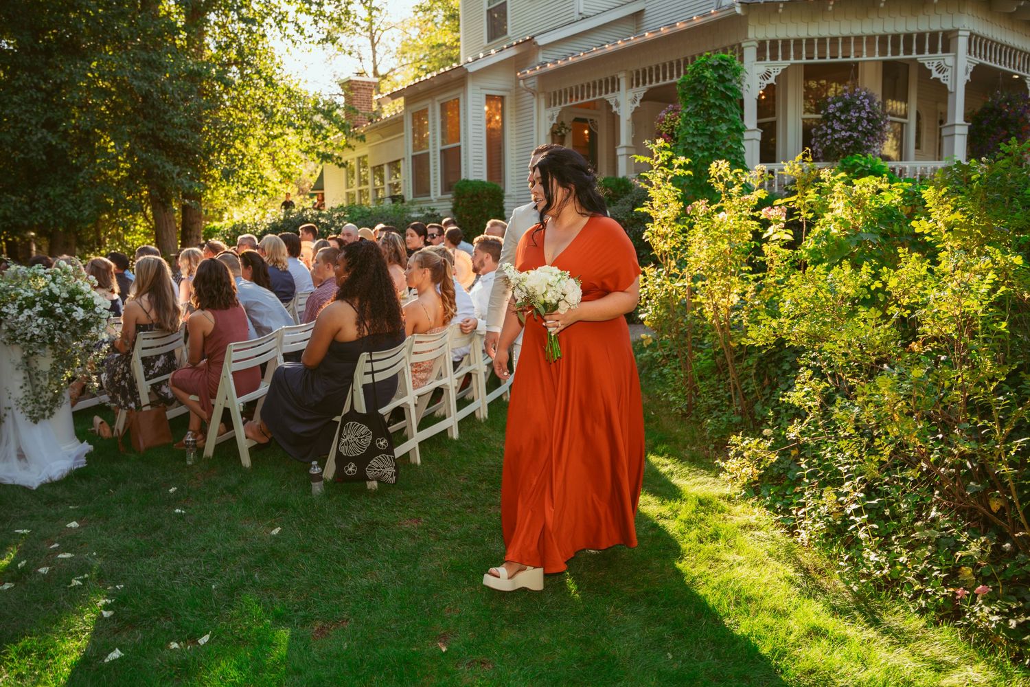 Wedding guests seated in white chairs on lawn with Victorian house in background.