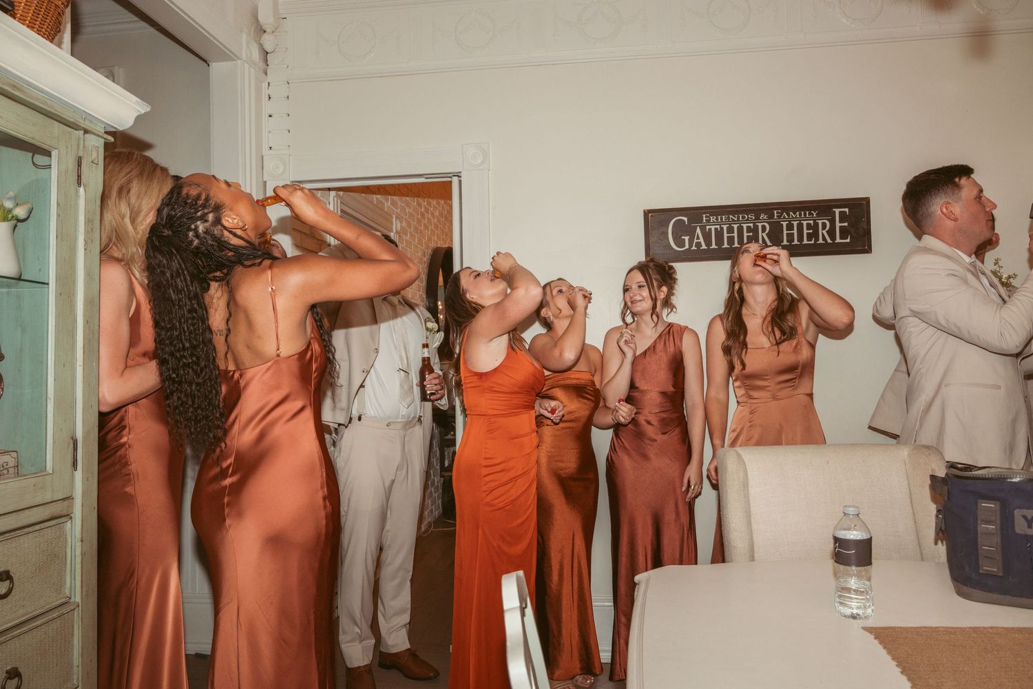 Wedding party members in coral dresses and suits pose together indoors for formal photos.