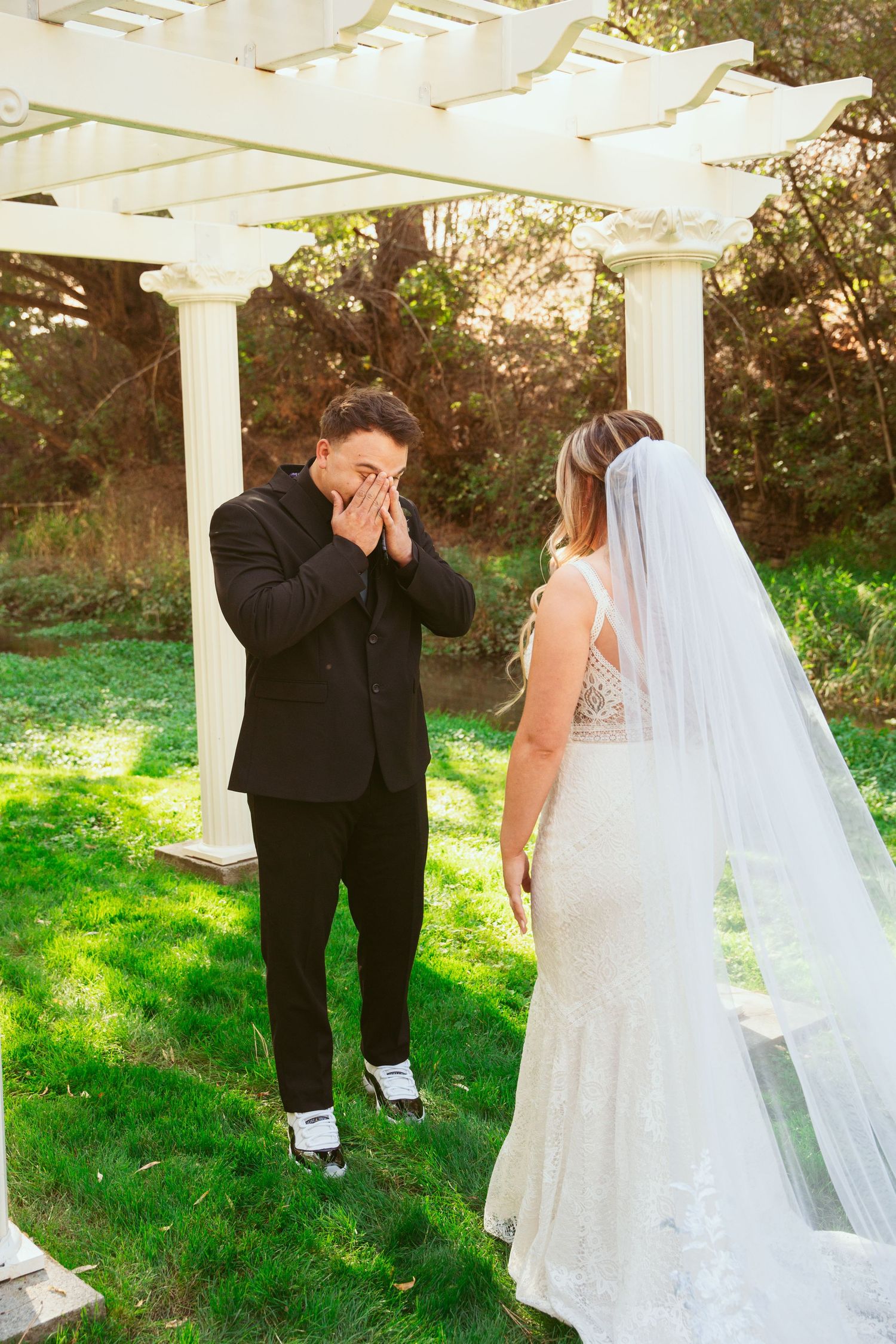 A romantic first look moment captured between a bride and groom under a white pergola in a garden setting.