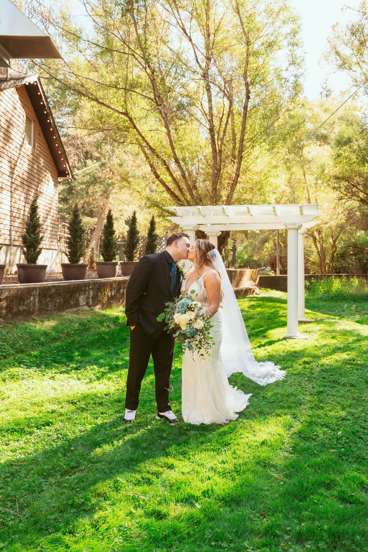 A wedding couple shares intimate moments under a white pergola on a sunny lawn.