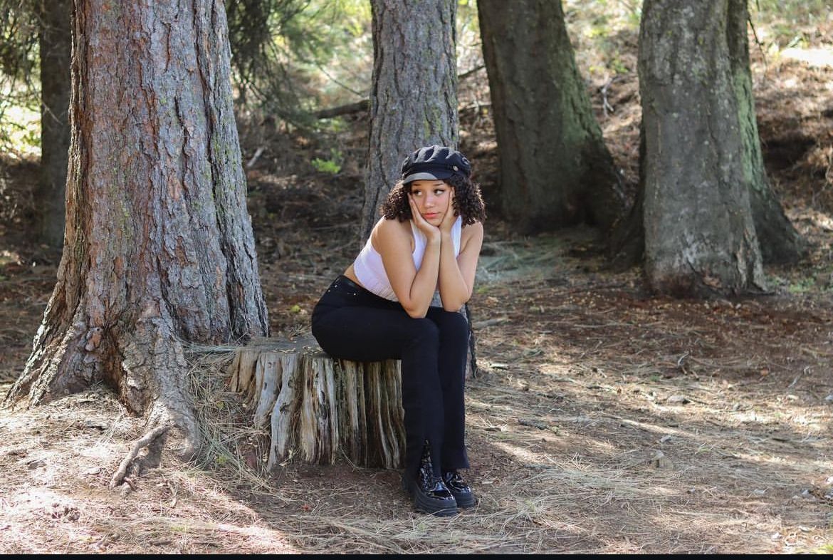 Someone crouches near a large tree trunk in a forest while wearing hiking gear and examining the ground.