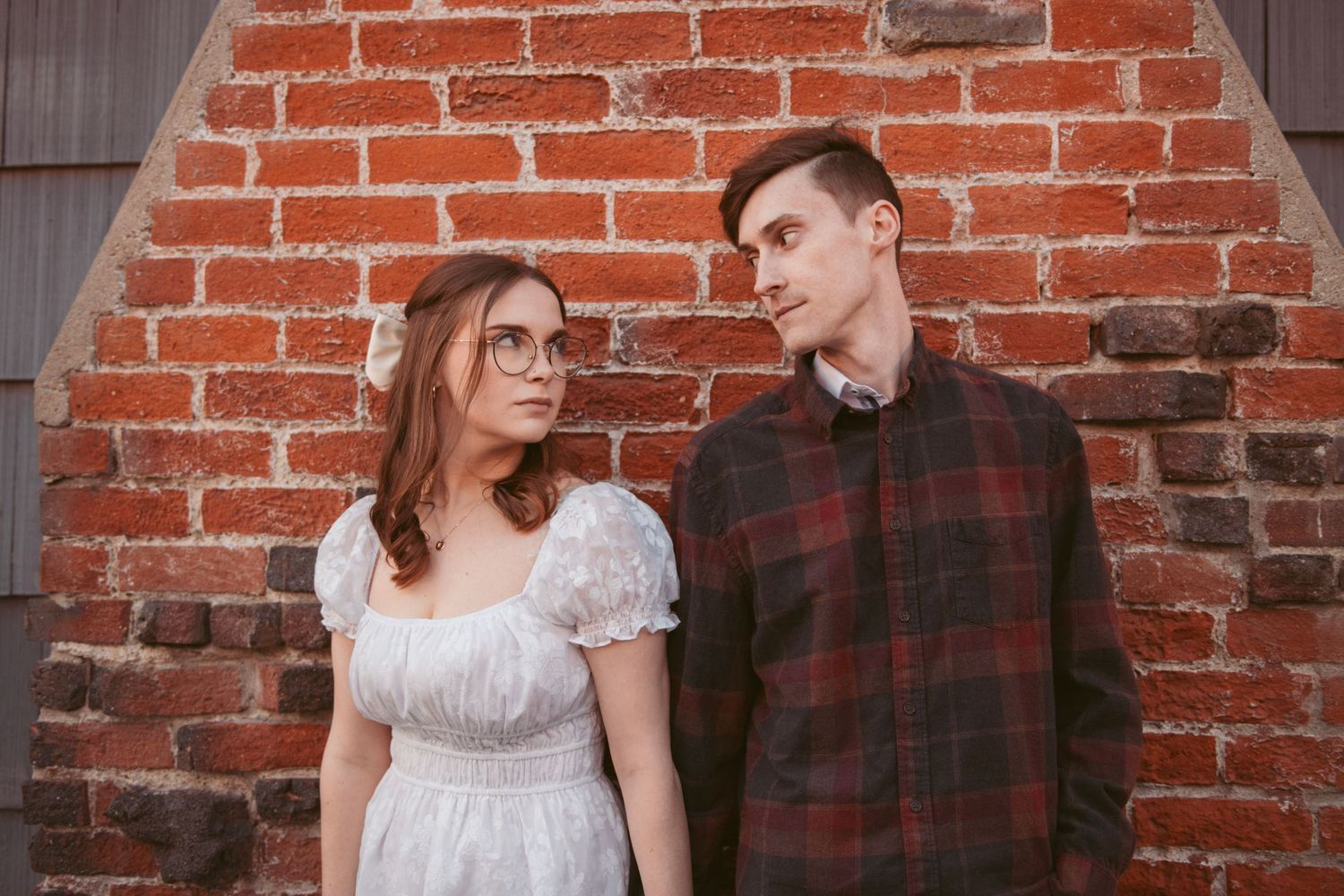 Couple in contrasting outfits posed against rustic brick wall.