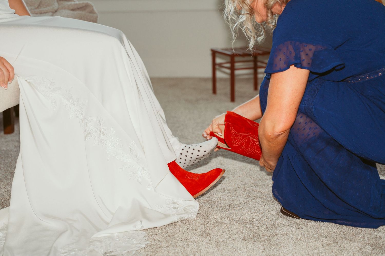 Wedding preparation scene showing red boots and dress hem on gray carpeted floor.