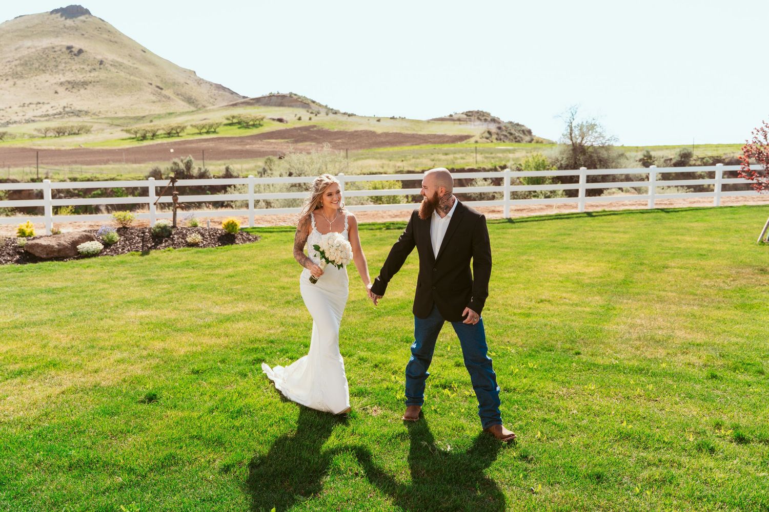 Wedding couple walking hand in hand across green field with mountain landscape.