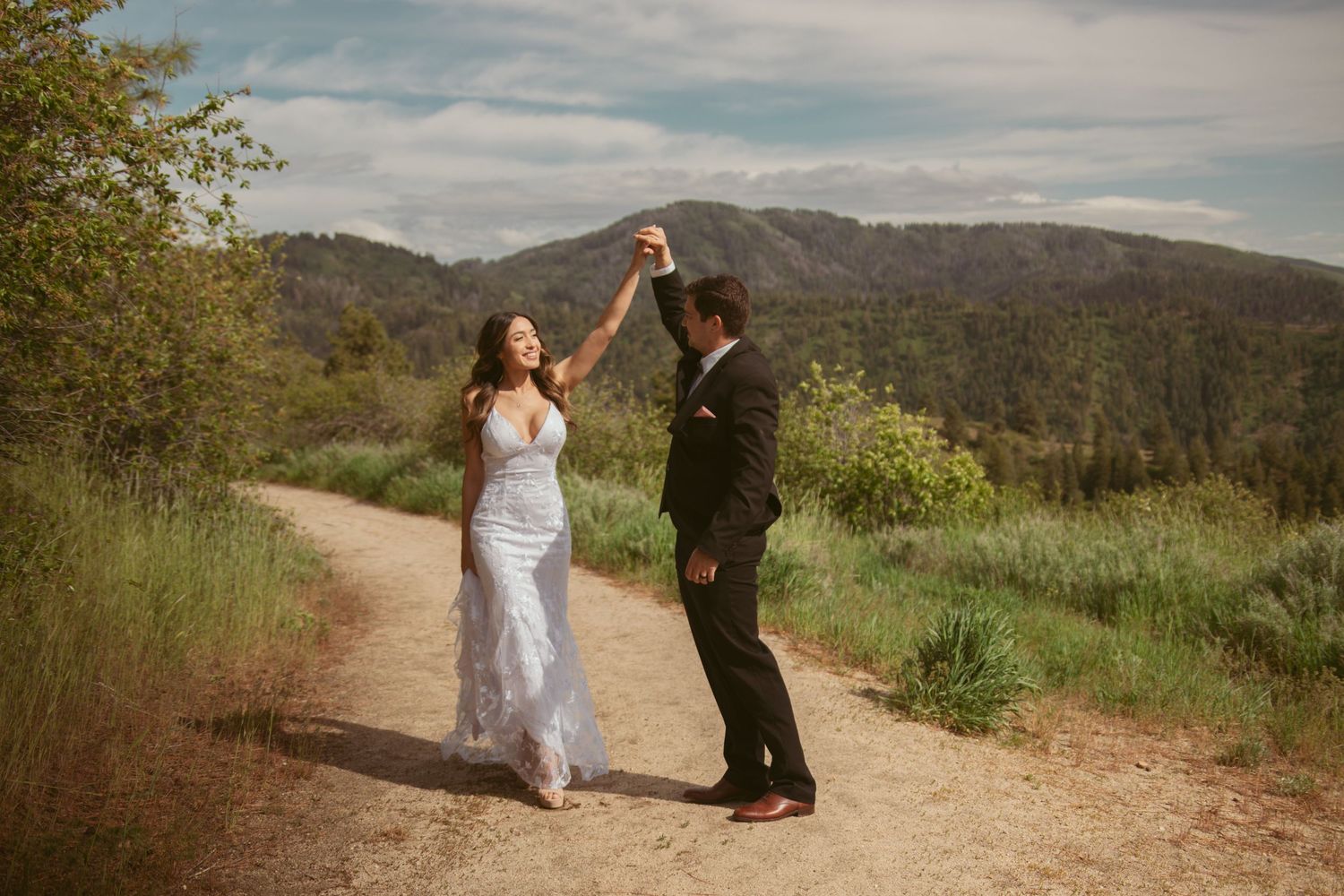 A couple dances together on a scenic mountain trail with blue skies and rolling hills in the background.
