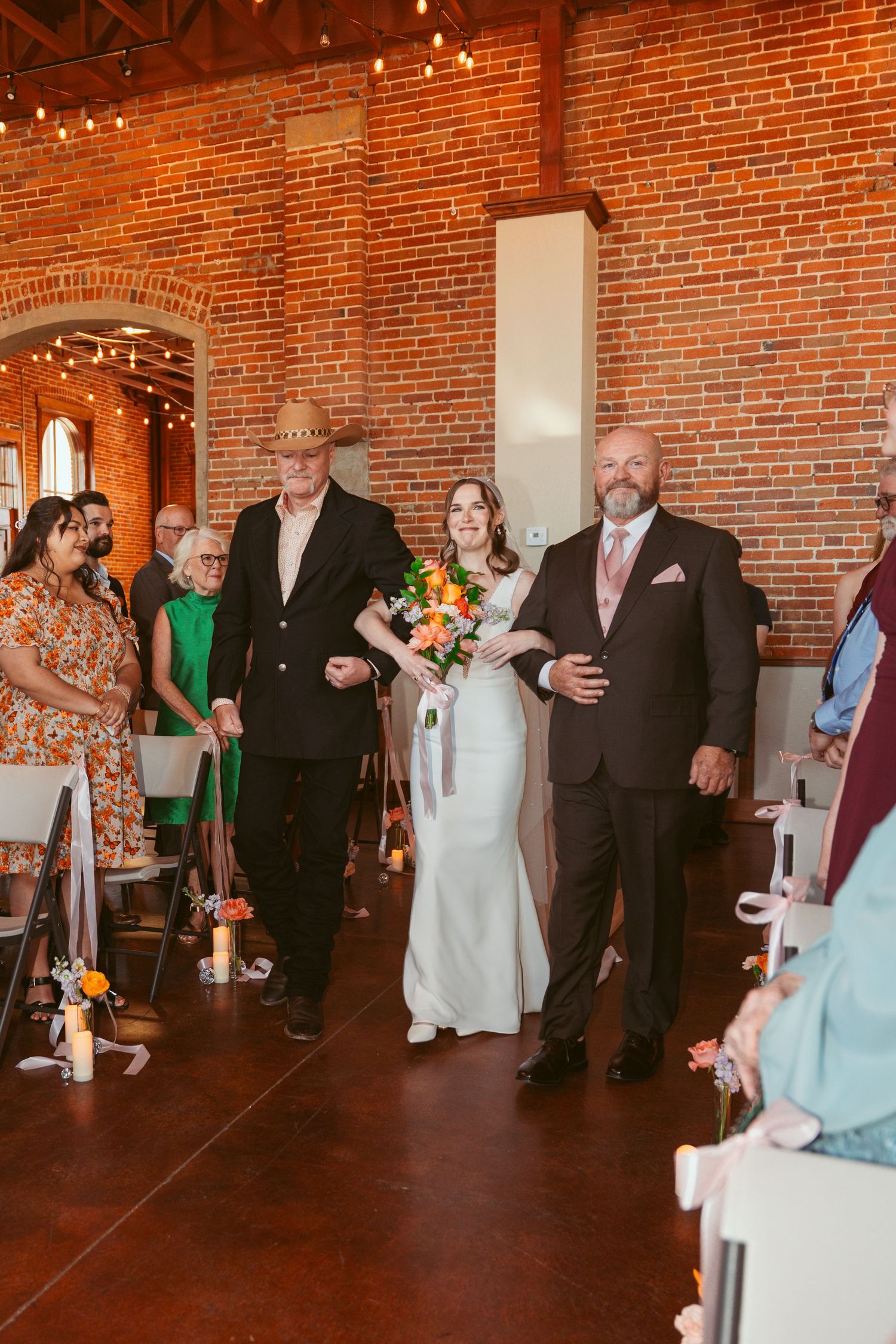 Wedding party members line up during ceremony in rustic brick venue with string lights.