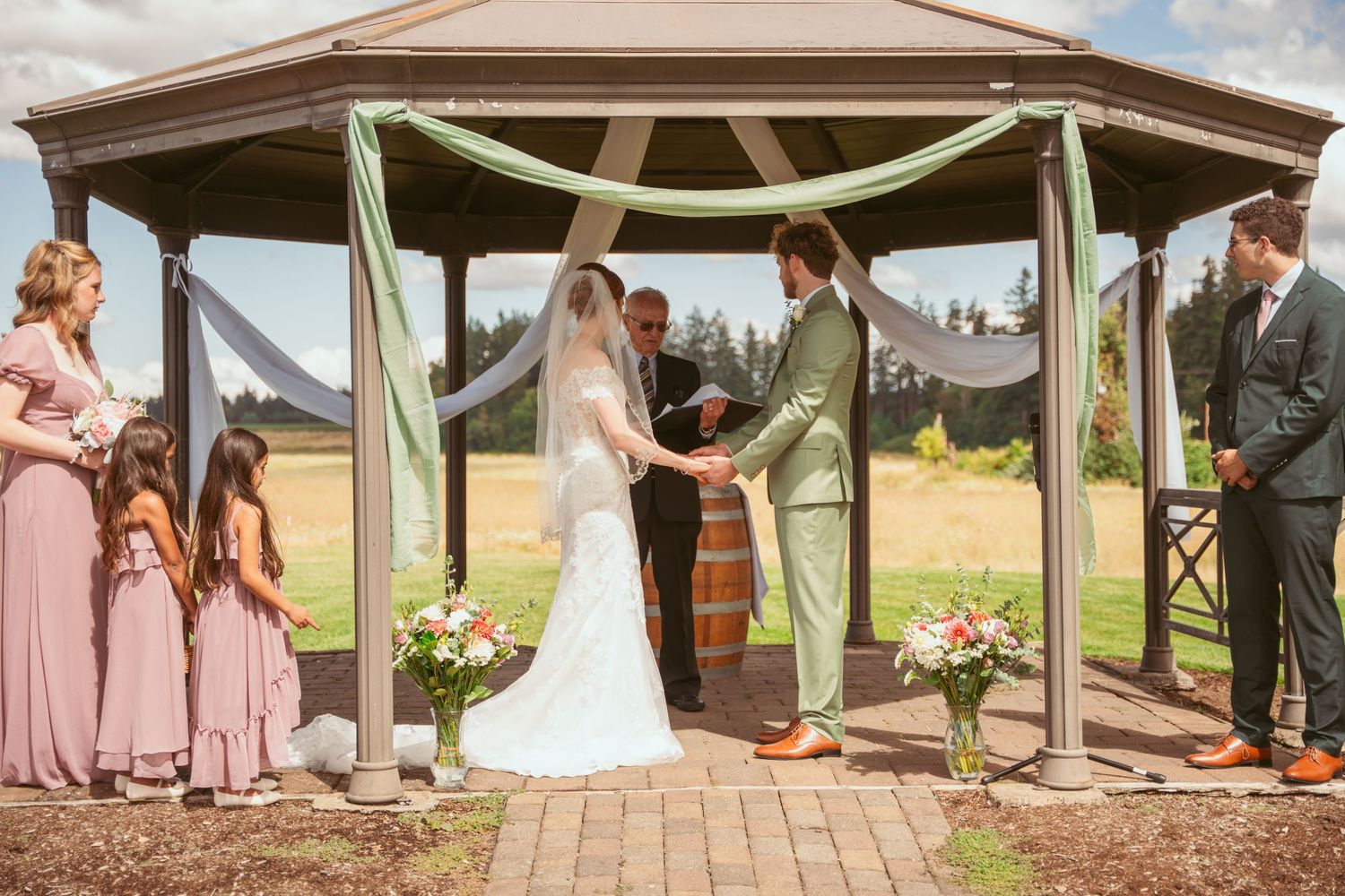 Wedding ceremony taking place under wooden gazebo with pink floral arrangements and draped fabric.