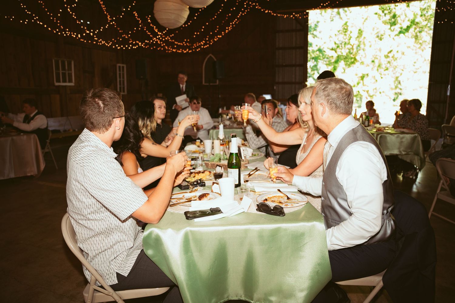 Long tables set for dinner with white tablecloths and string lights overhead in rustic barn venue.