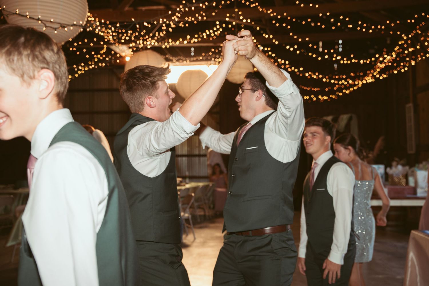 Wedding party members join hands in celebration under twinkling lights at a barn reception.