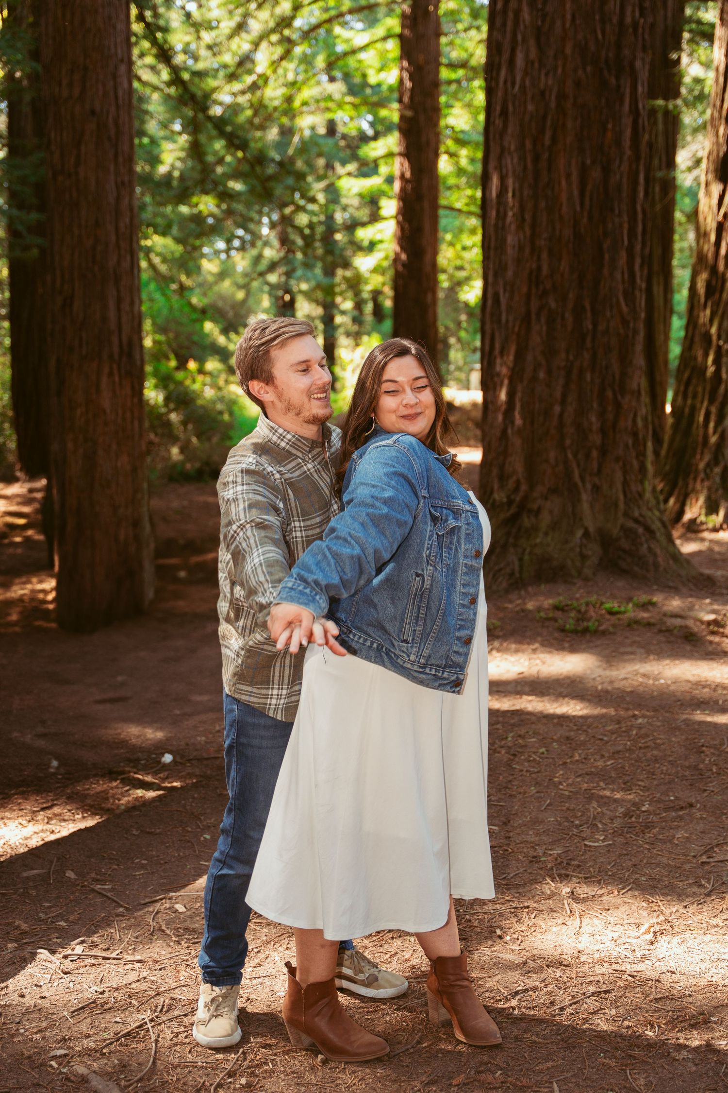 Two people share a joyful dance-like moment surrounded by towering redwood trees.