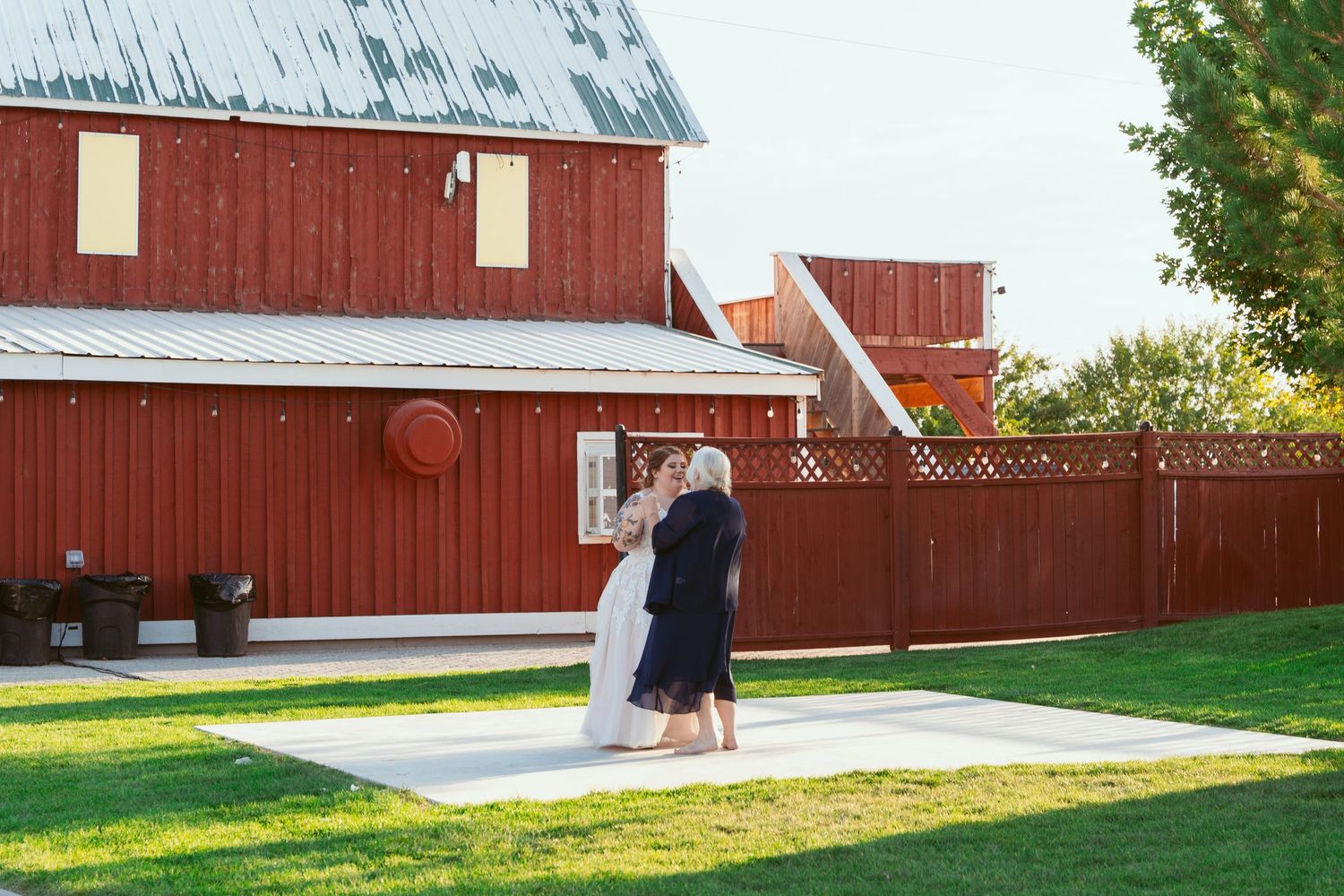 Couple dancing on concrete pad outside red barn venue on sunny day.
