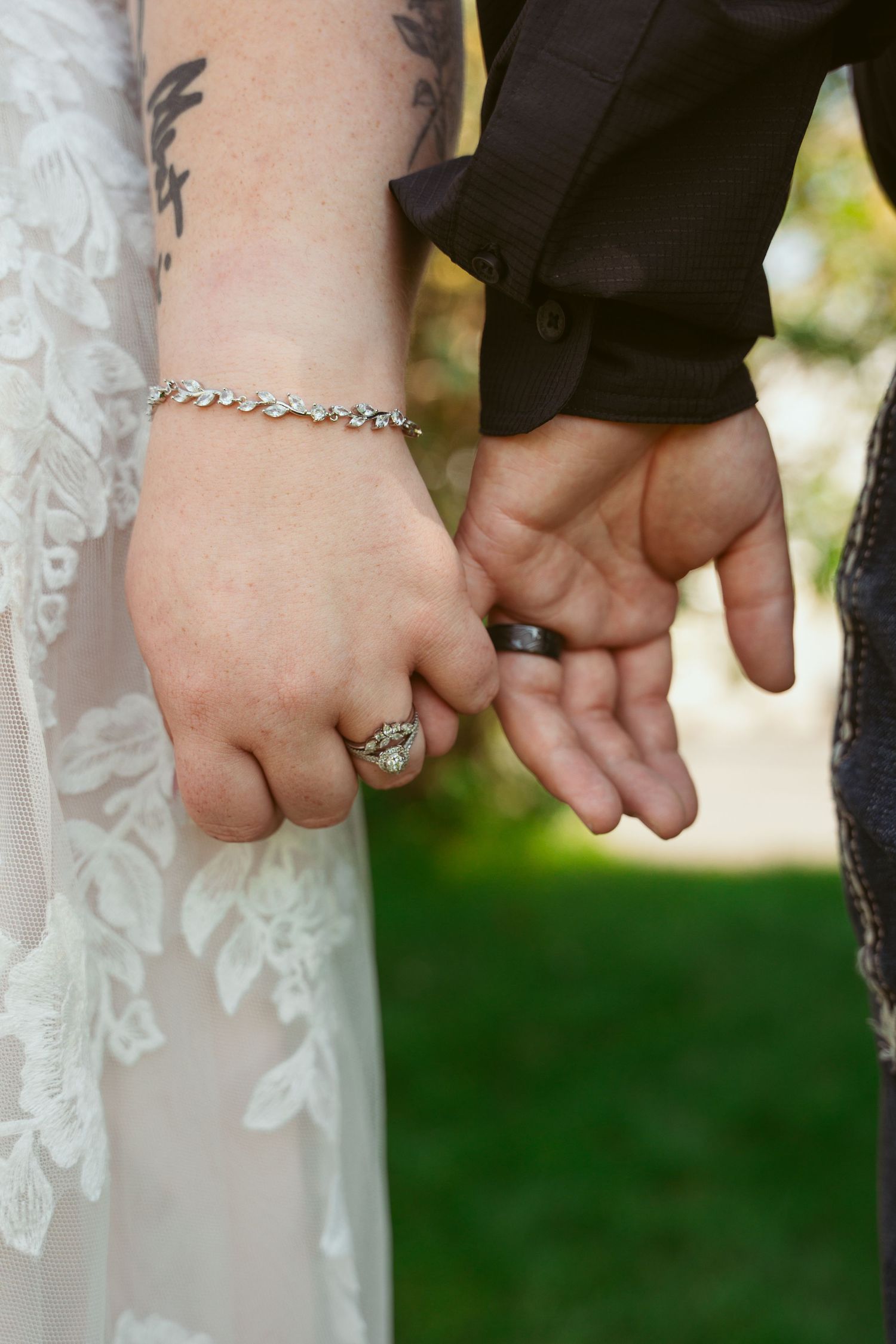 Close-up of couple holding hands showing wedding rings and a delicate bracelet against a blurred green background.