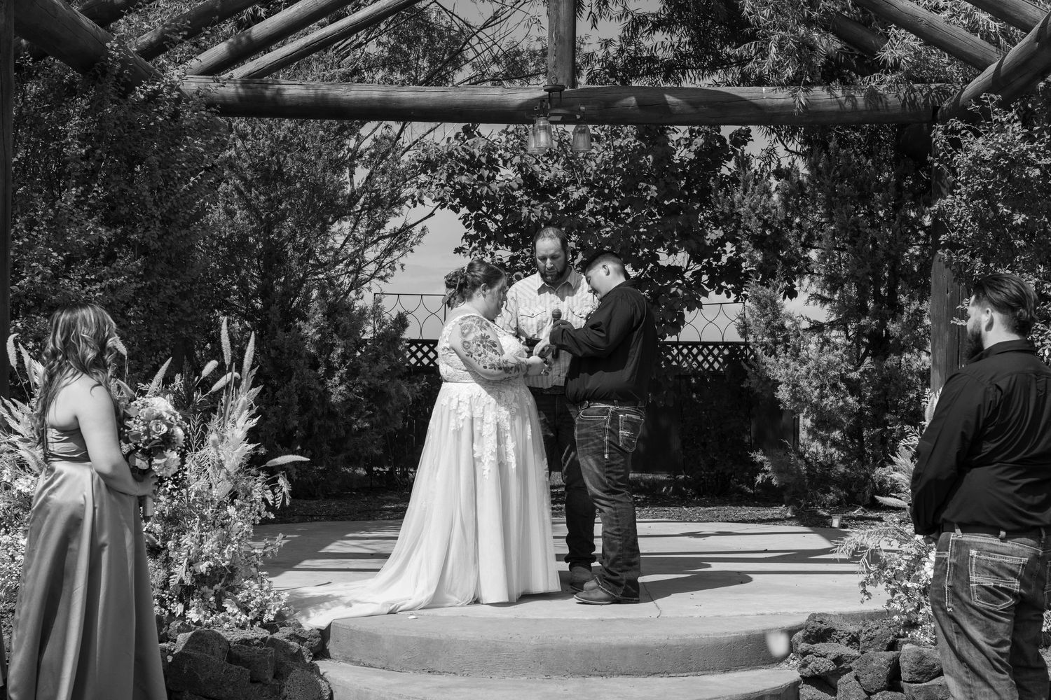 Black and white image of outdoor wedding ceremony with guests seated under tree canopy.