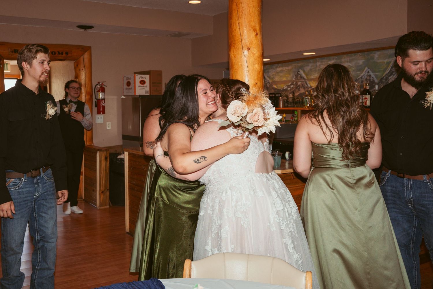 Wedding party members in olive green dresses and formal attire dance together at a reception venue with wooden decor.