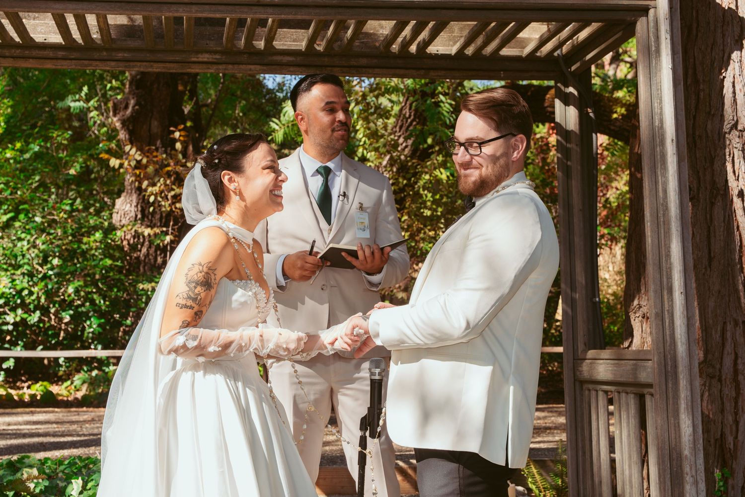 A wedding ceremony takes place under a wooden gazebo with greenery in the background during a sunny day.