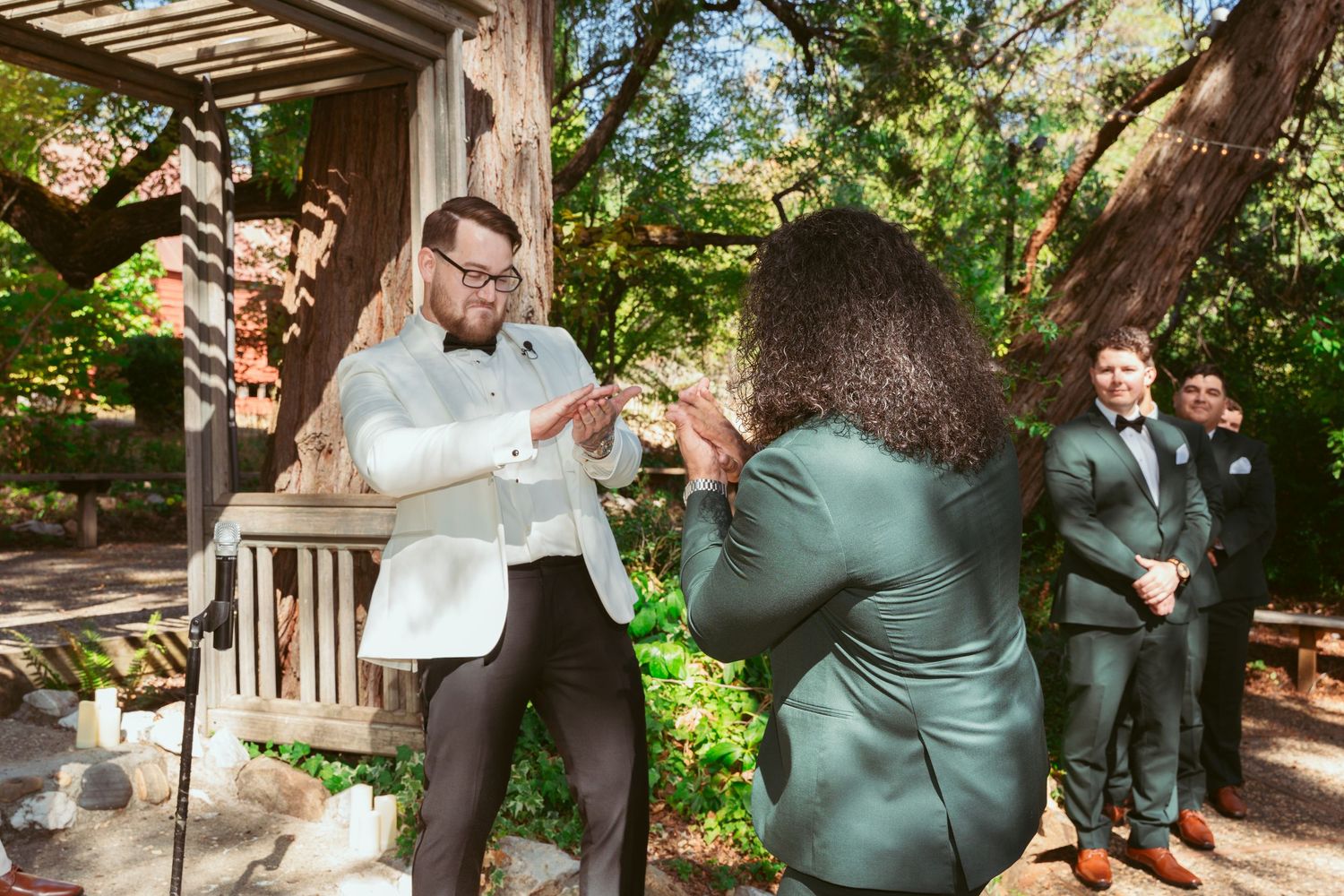 Romantic wedding couple embraces in a lush garden setting with rustic wooden structures.