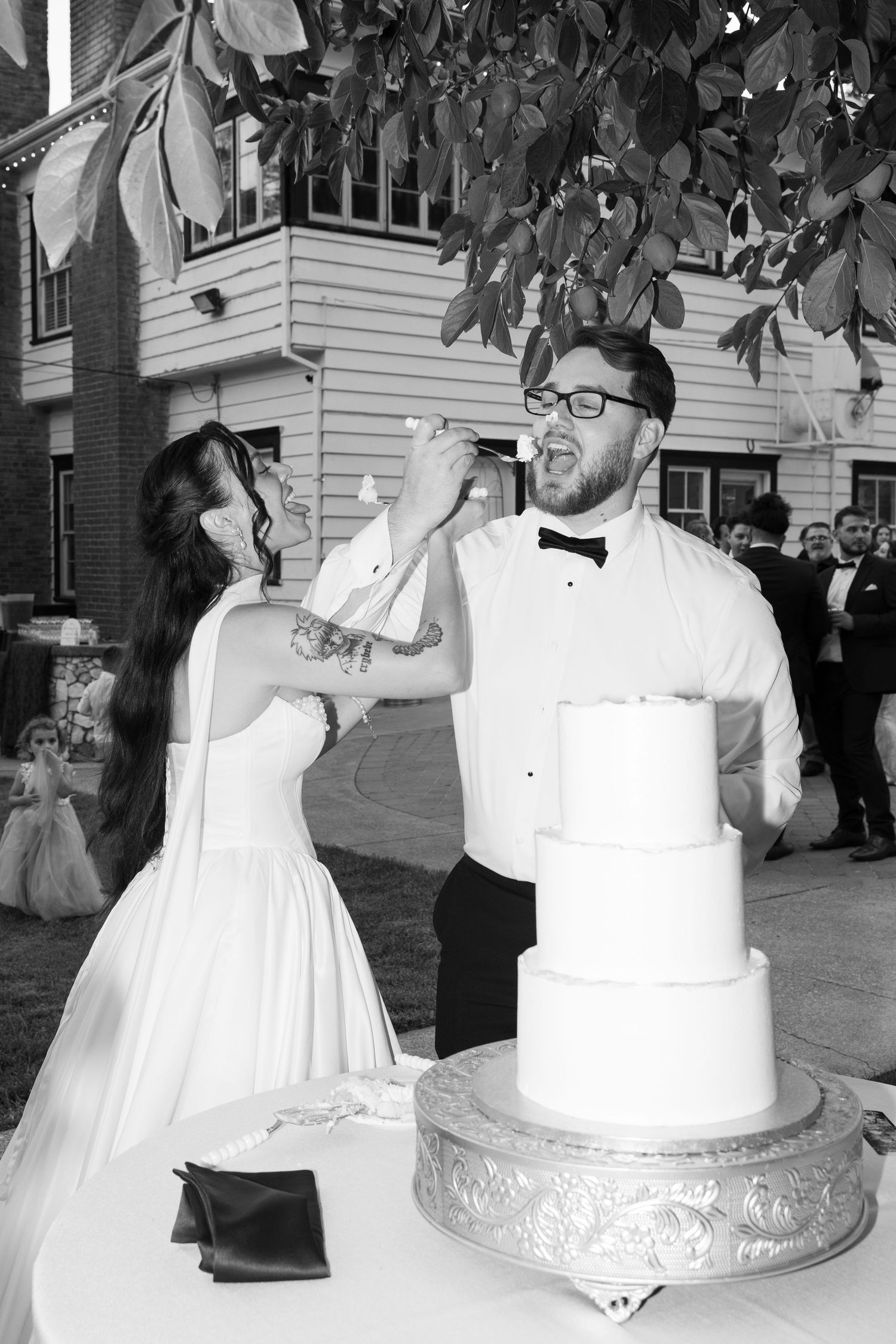 A couple shares a playful moment while cutting their elegant white wedding cake at an outdoor reception.
