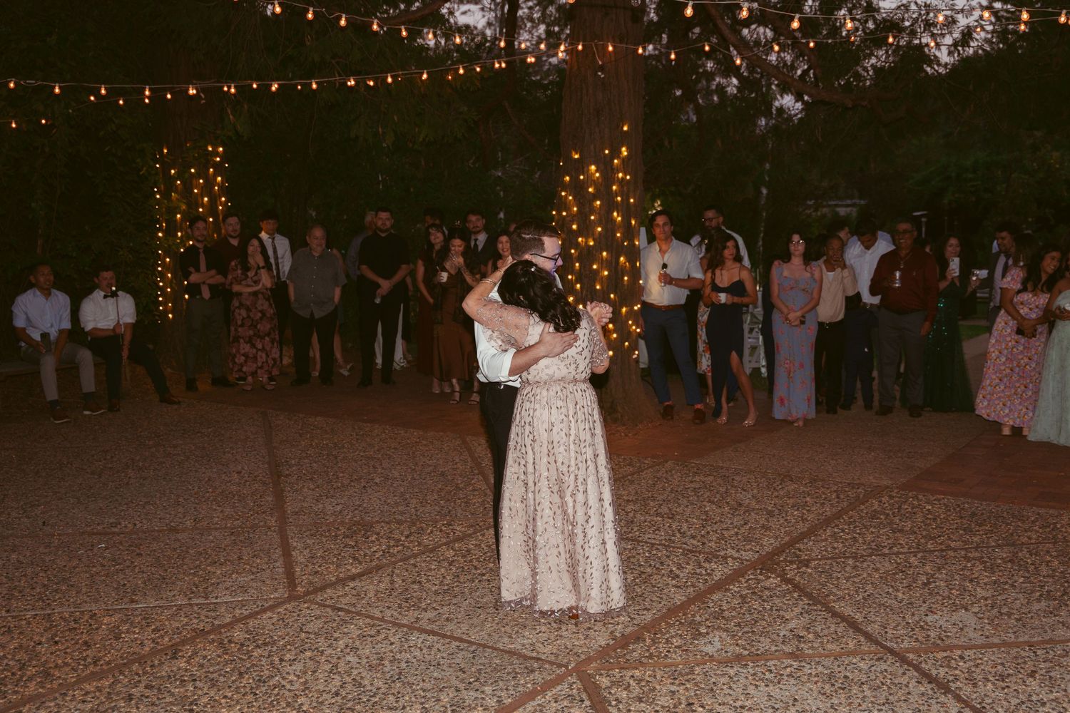 A couple dances intimately on an outdoor patio illuminated by string lights while guests look on.