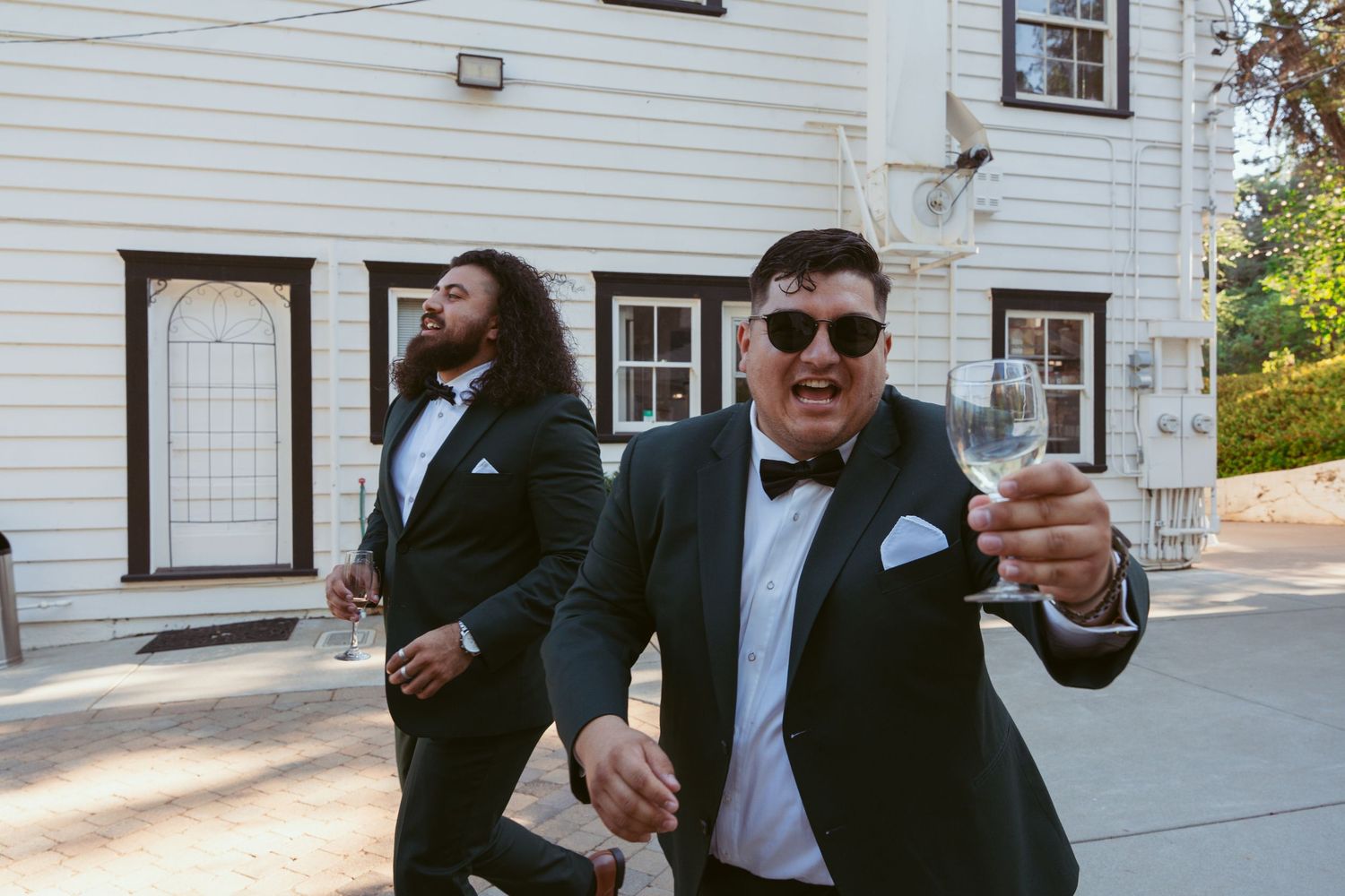Groomsmen in formal attire celebrate outside a white building during wedding festivities.