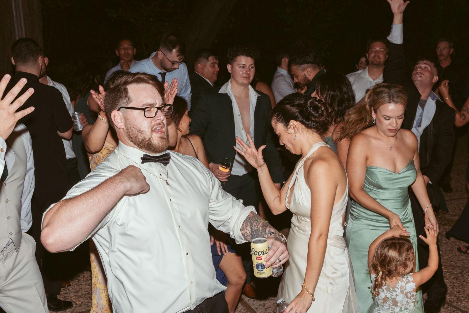 Wedding guests dancing energetically at a reception with people in formal attire on a dark dance floor.