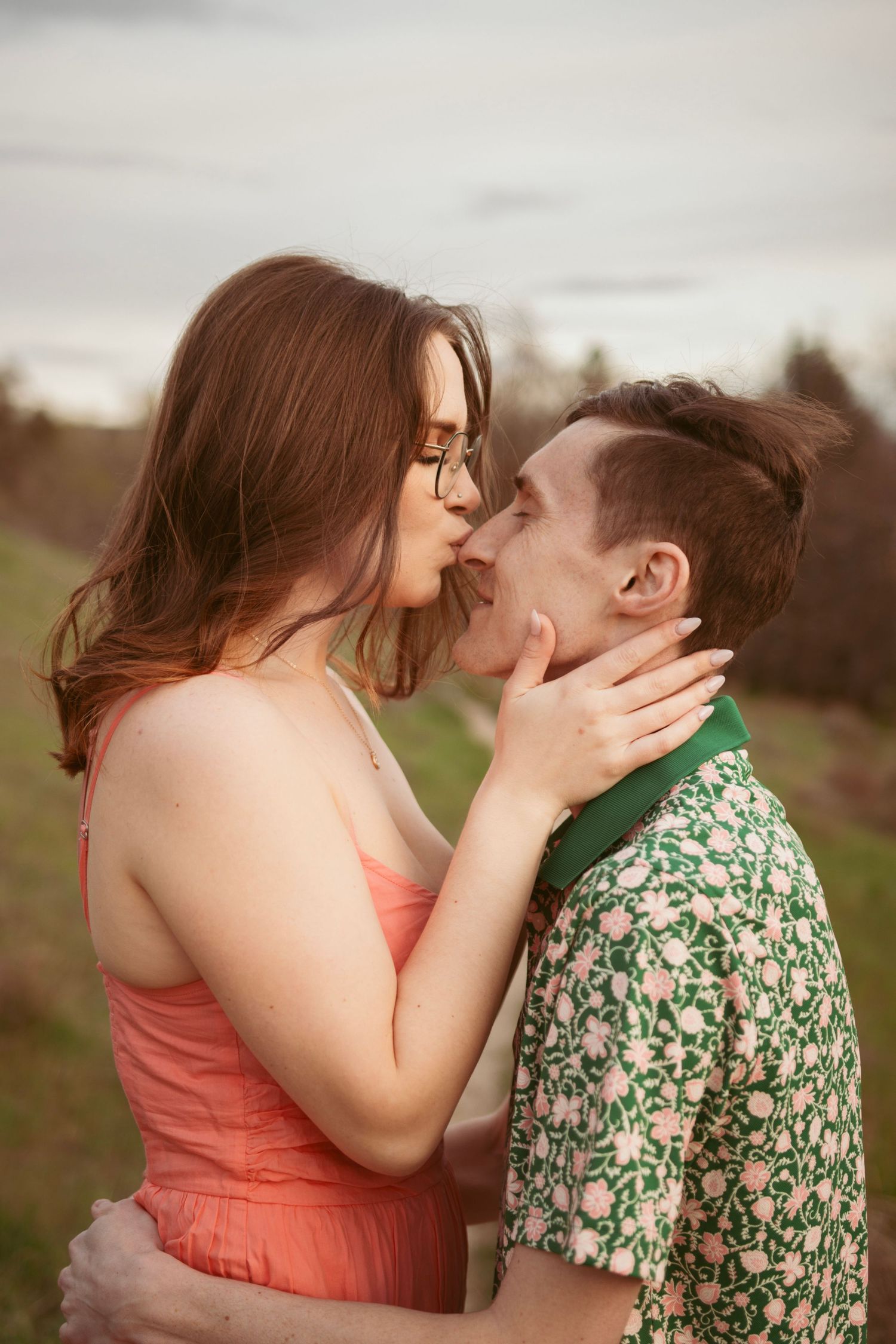 An intimate moment between a couple in a coral dress and floral shirt against a cloudy sky.