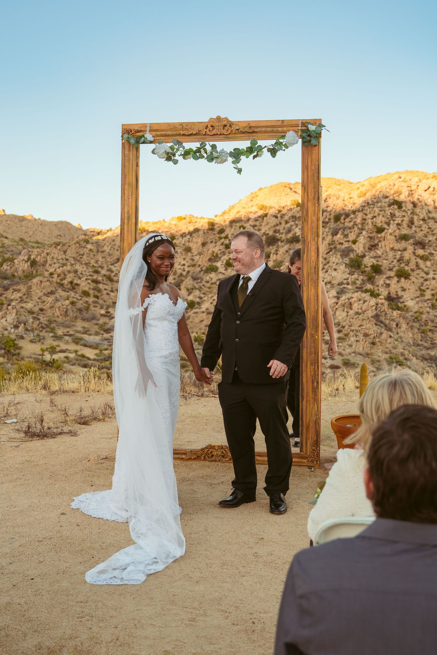 Desert wedding ceremony with couple holding hands under wooden arch as guests look on.
