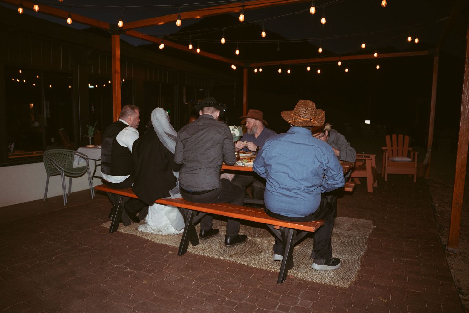 String lights illuminate a rustic outdoor reception area with wooden benches at dusk.
