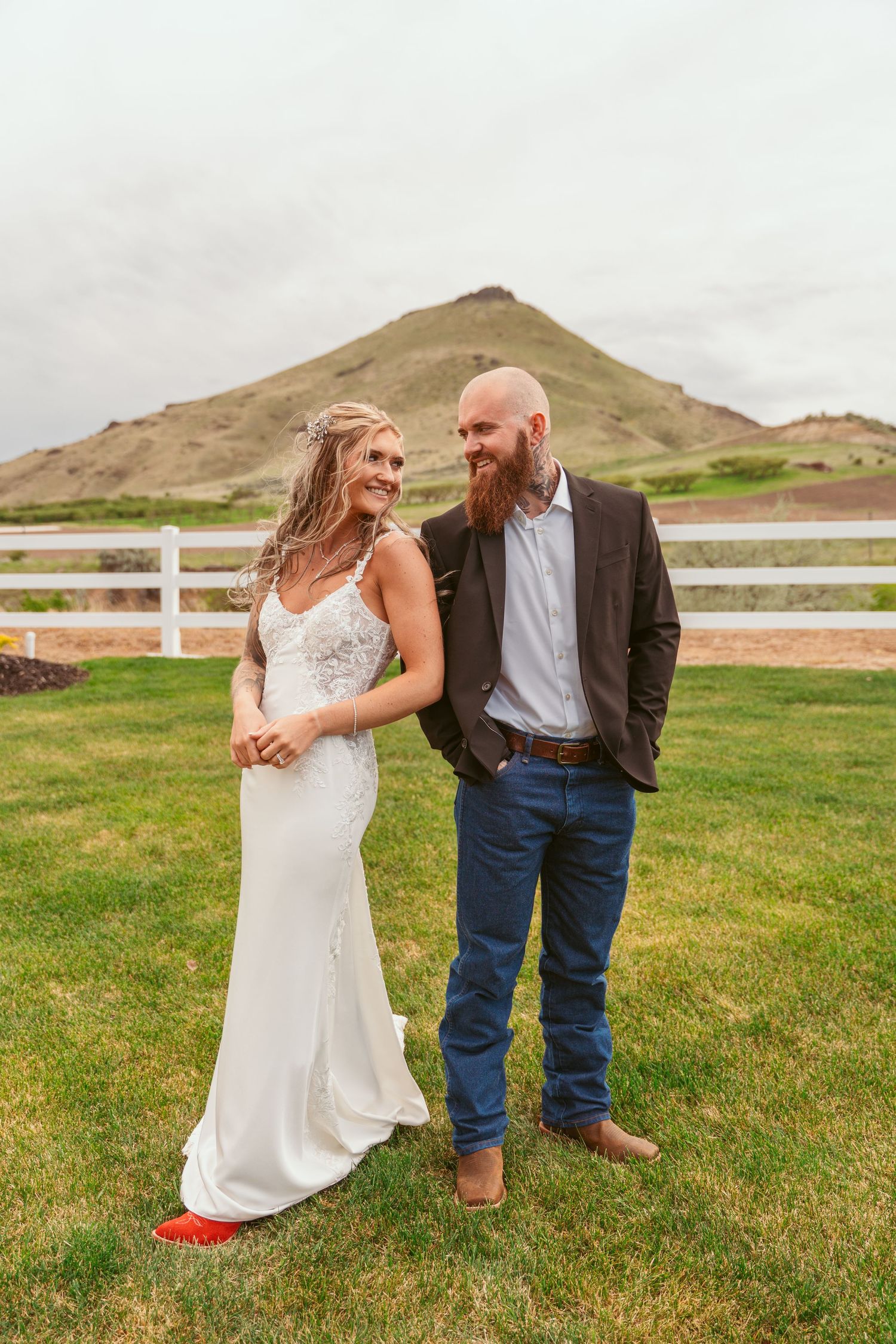 Couple in wedding attire standing on grass with desert mountain in background.