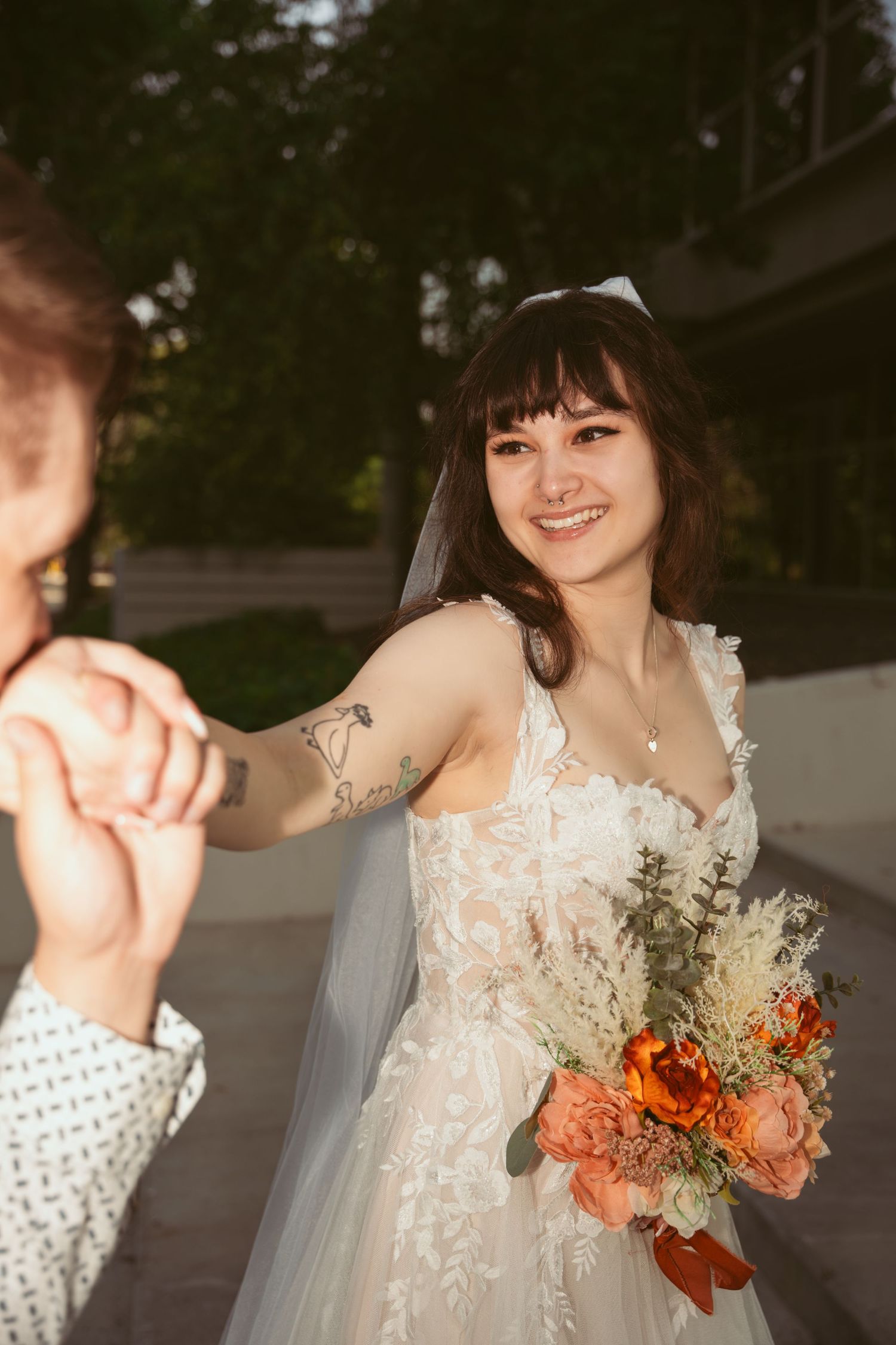 A bride in a lace dress holds a bouquet of orange flowers during an outdoor photoshoot.