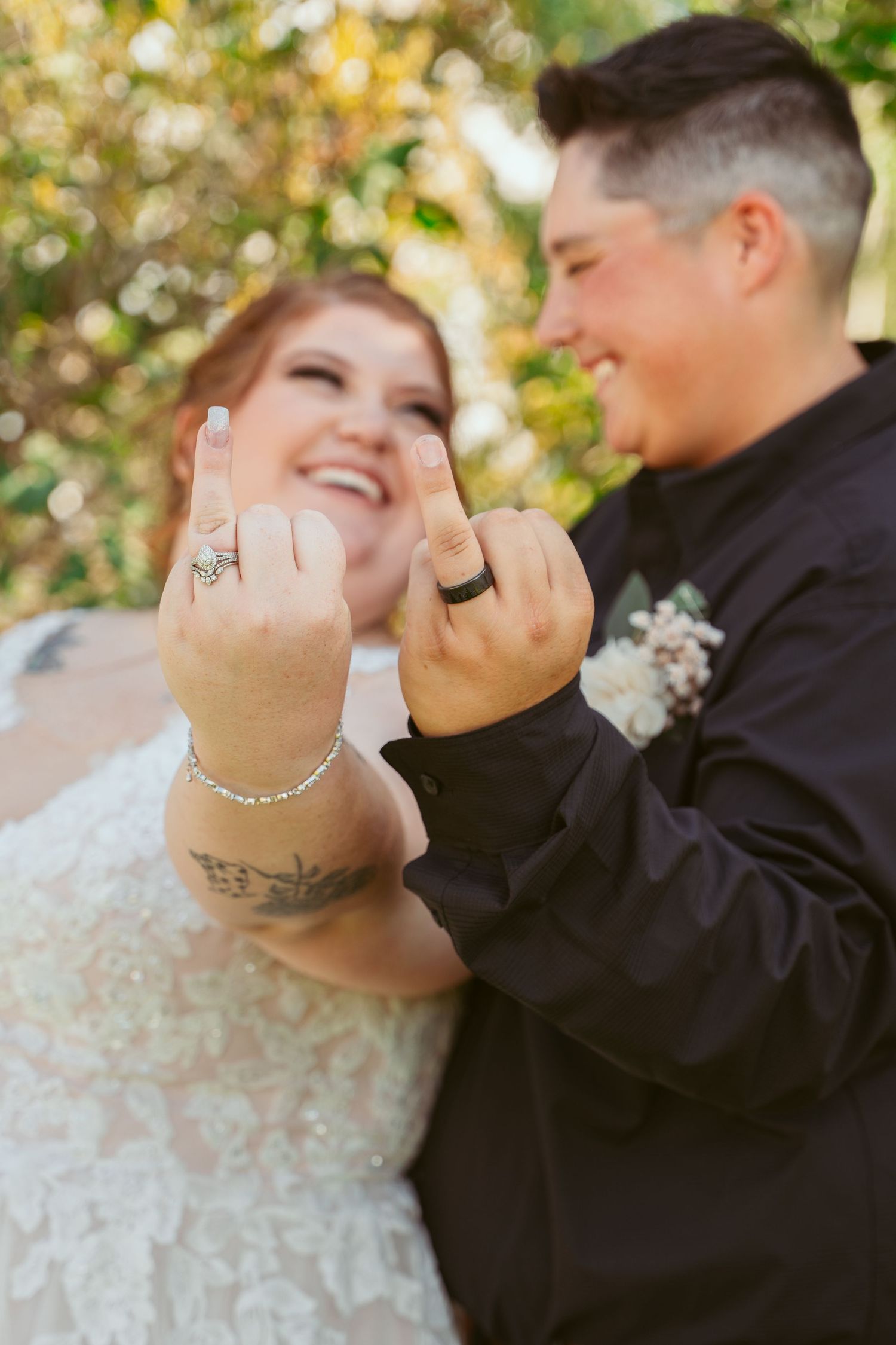 A joyful couple in wedding attire showing off their rings and laughing together outdoors.