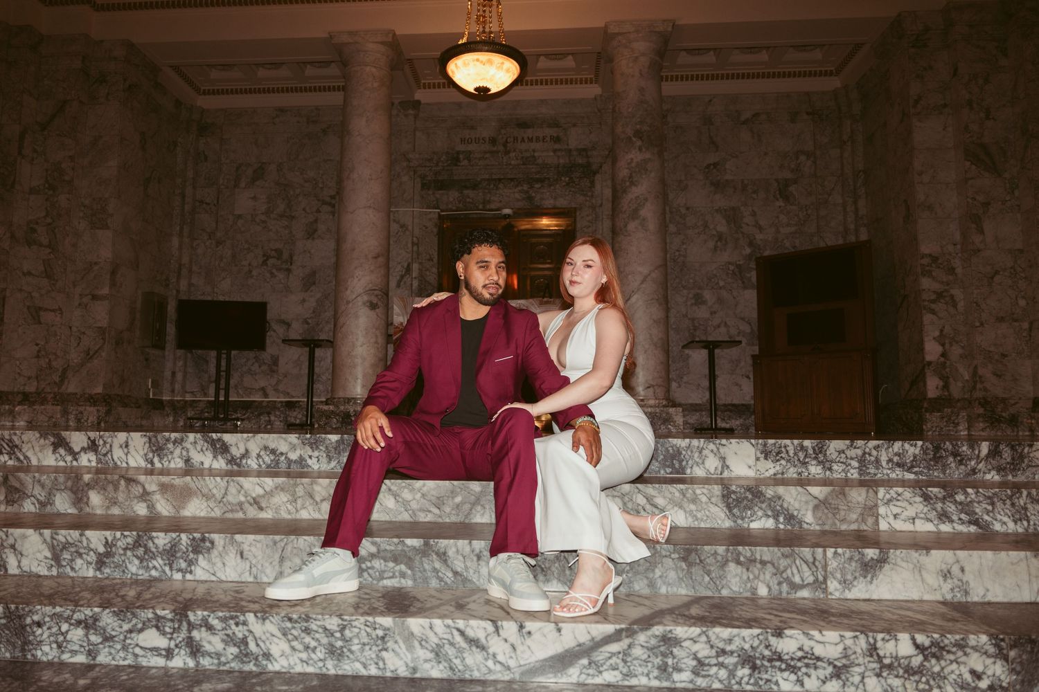 A couple in coordinated formal wear sits casually on marble steps in an elegant building.
