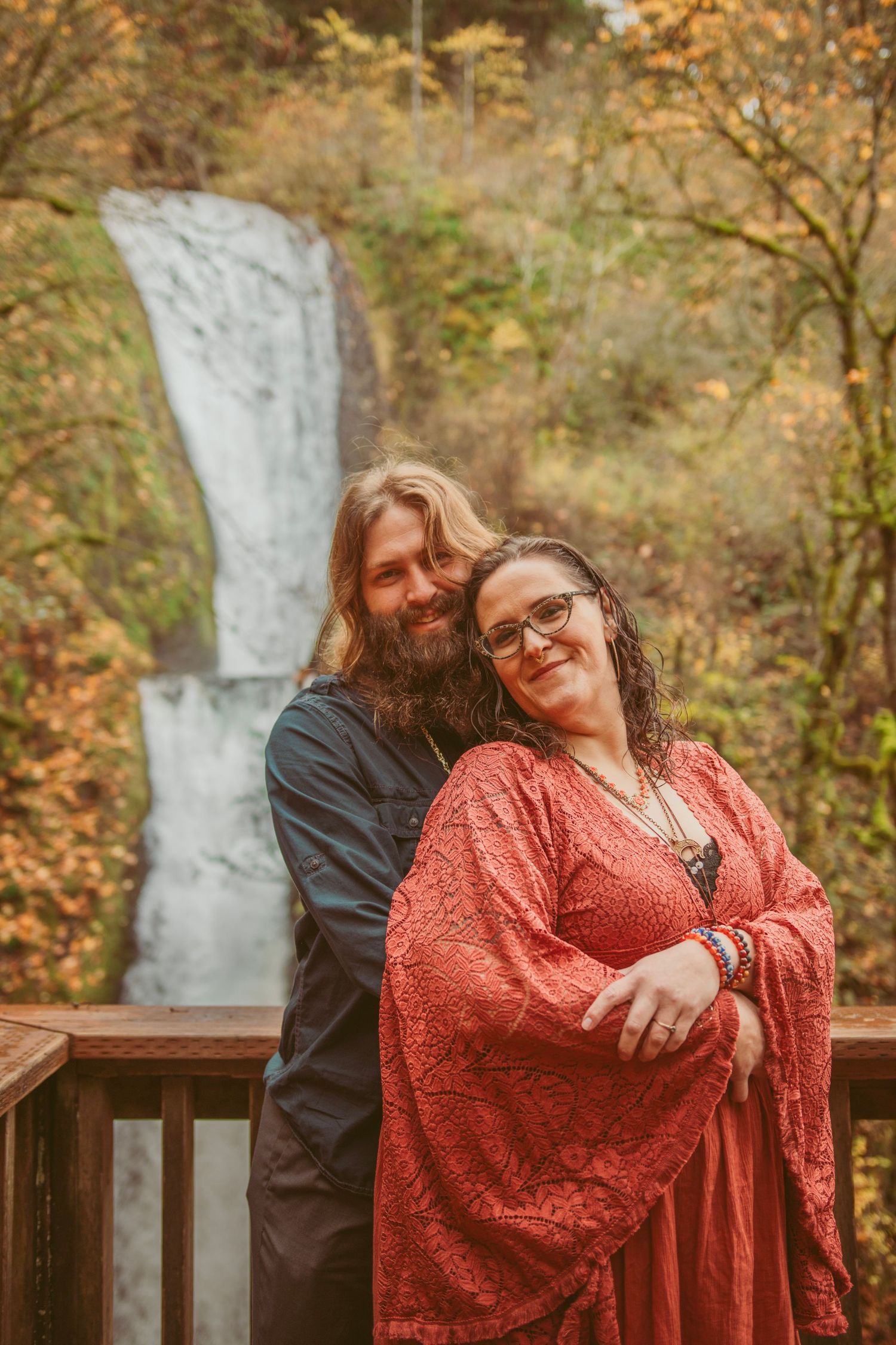 Romantic photoshoot on wooden deck with waterfall and autumn foliage in background.