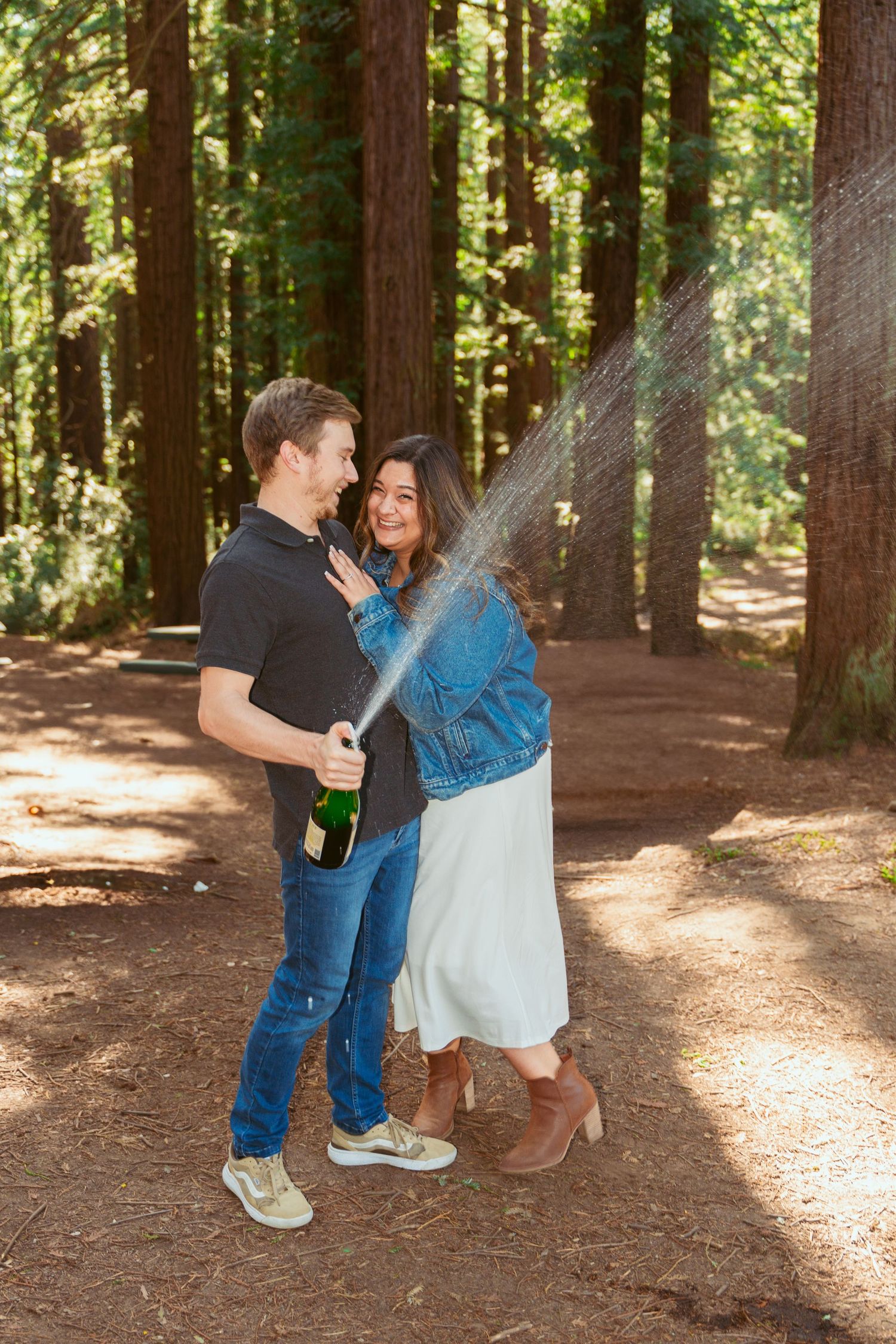 A couple celebrates with champagne in a sunlit redwood forest.