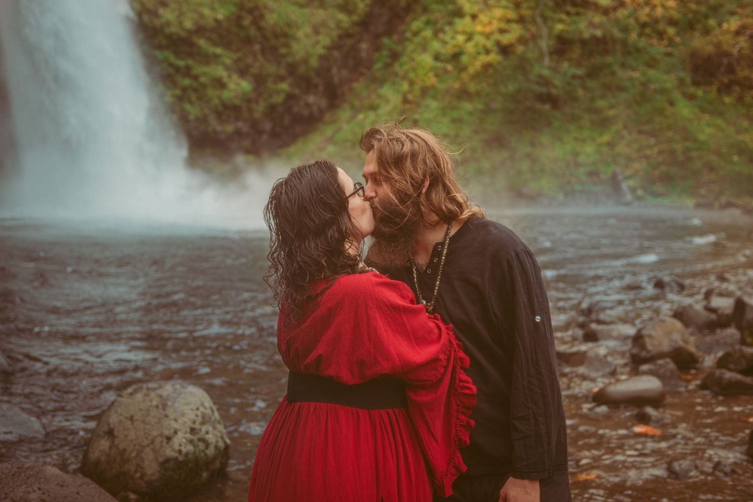 Artistic photoshoot near a waterfall features dramatic poses and a striking red dress.